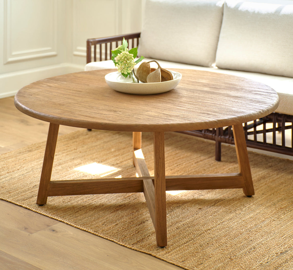 This is a lifestyle image of Aksel Coffee Table Ø120 cm in teak indoor. With a bowl of flowers before a rattan sofa.