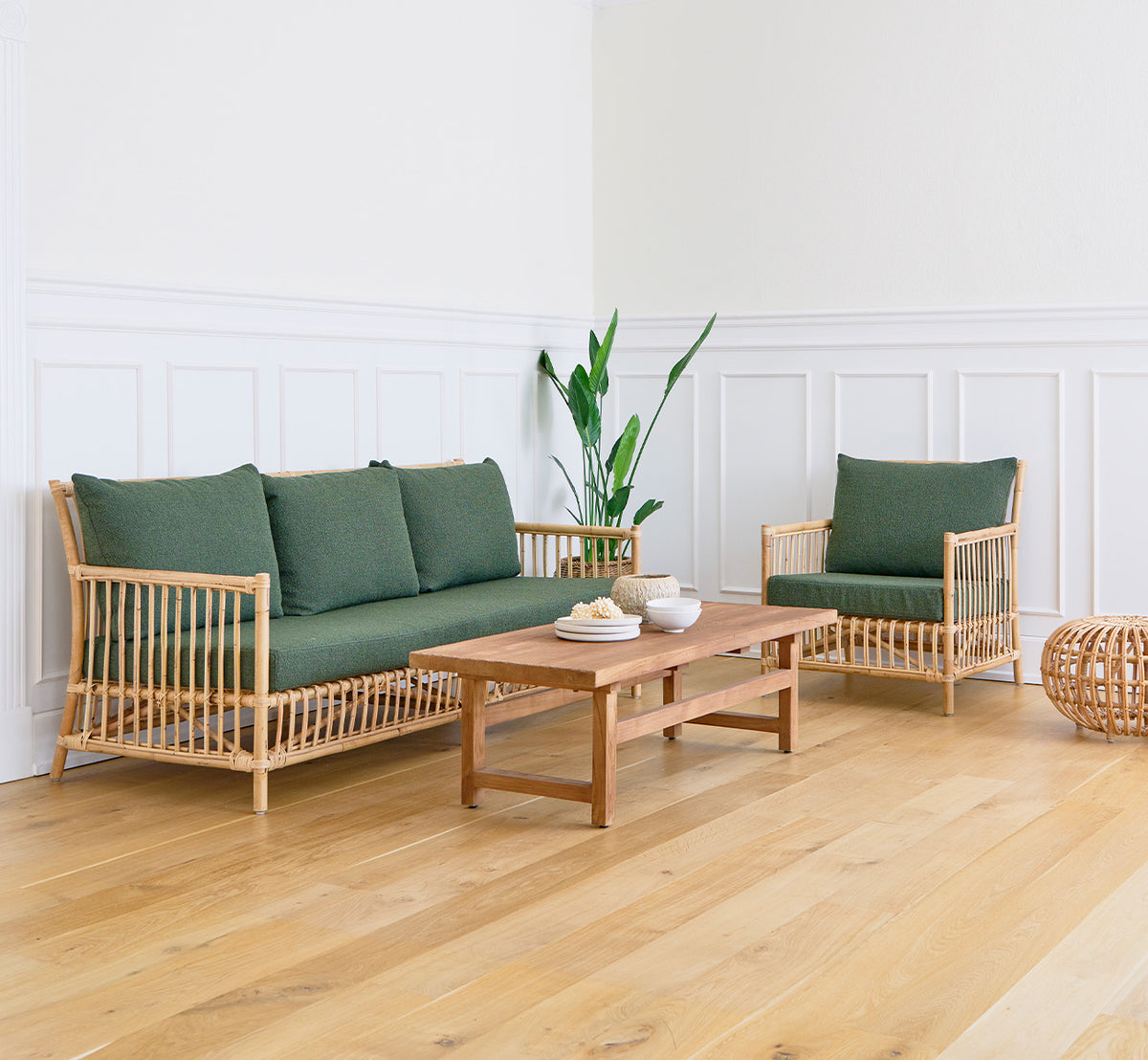 This is a lifestyle image of Alfred Coffee Table in teak indoor, in a bright living room with white panelled walls and a green-cushioned sofa.