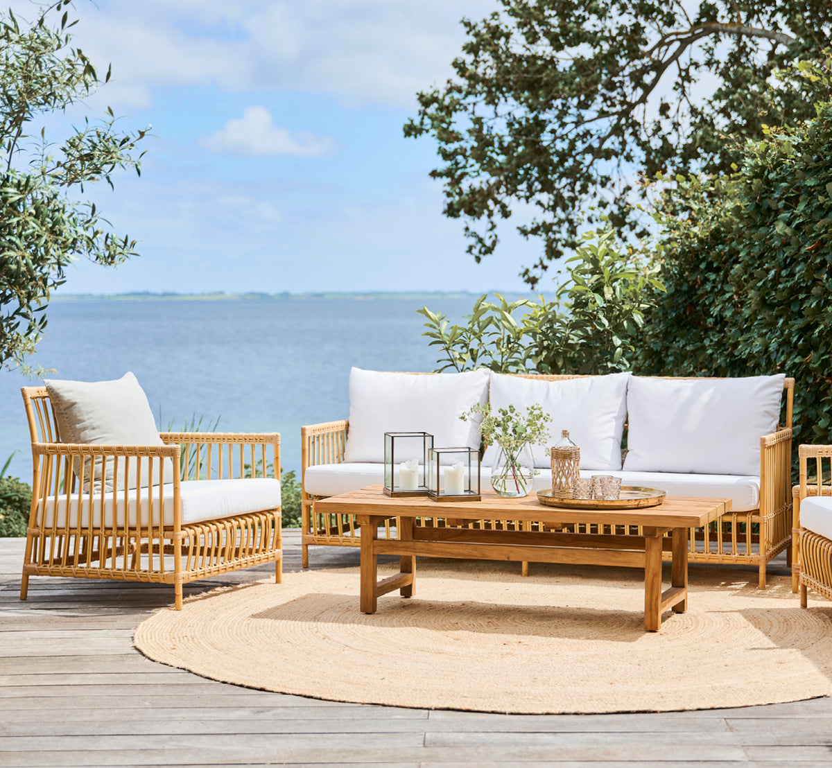 This is a lifestyle image of Julian Teak Lounge Table 140x55 cm in teak outdoor. On a terrace with lounge furniture, a jute rug, and ocean views.