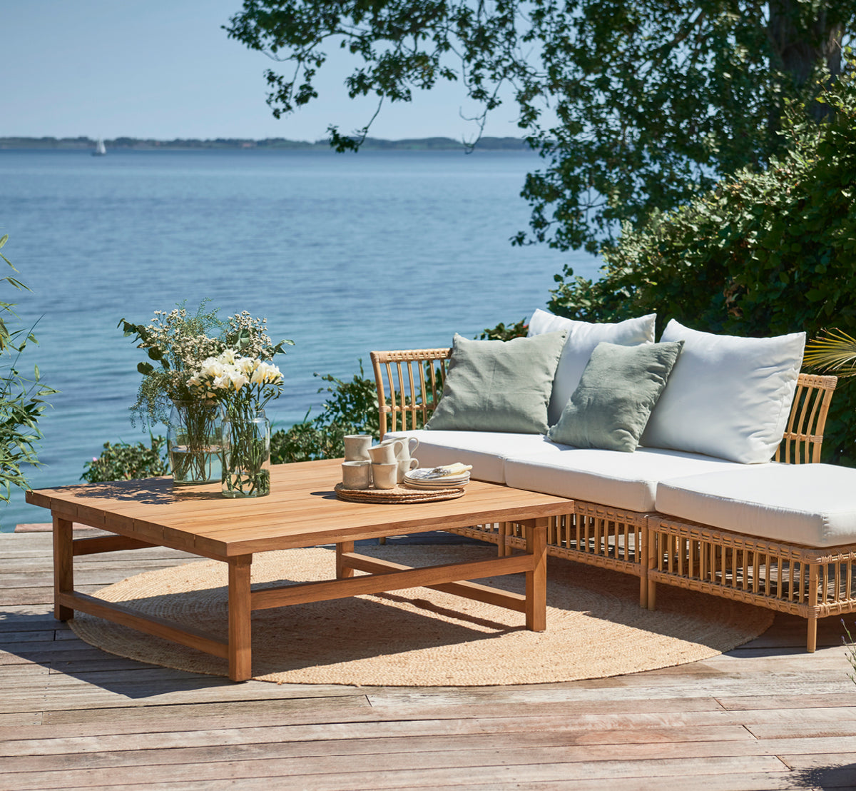 This is a lifestyle image of Julian Teak Lounge Table 120x120 cm in teak outdoor. On a seaside deck with a lounge sofa, cushions, and a tea setting.