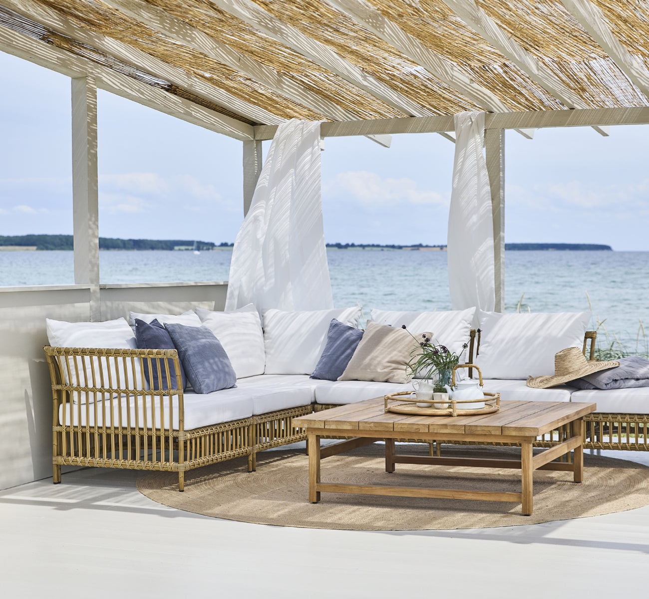 This is a lifestyle image of Julian Teak Lounge Table 120x120 cm in teak outdoor. Under a beach pergola with lounge seating and ocean views.