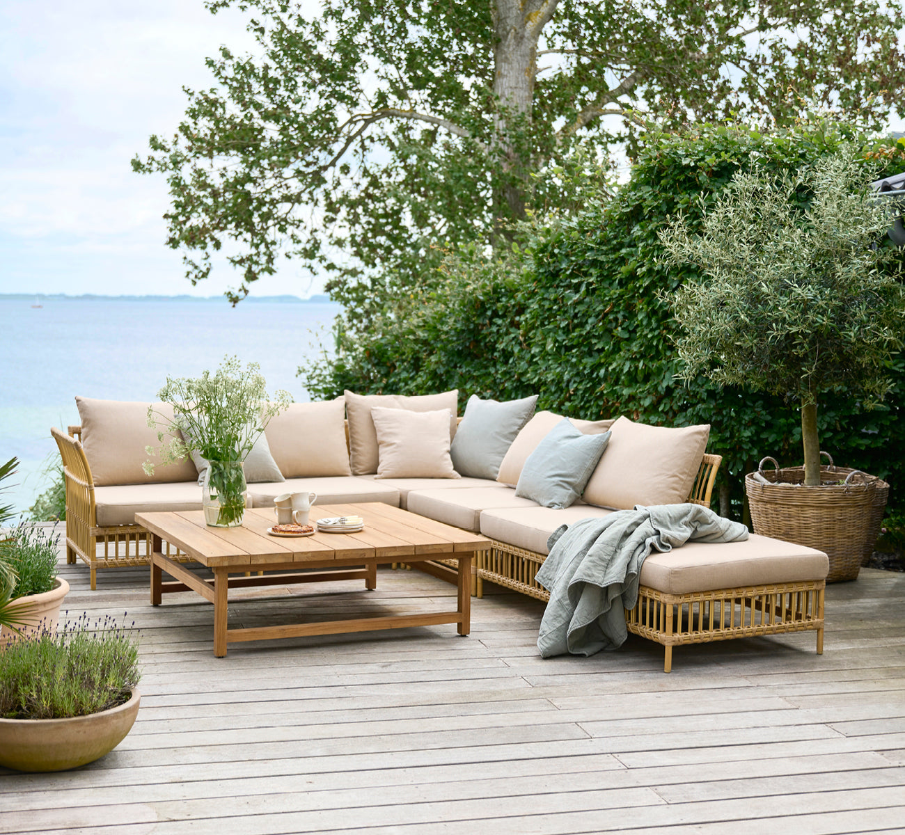This is a lifestyle image of Julian Teak Lounge Table 120x120 cm in teak outdoor. On a garden deck by the sea with lounge seating and lush greenery.