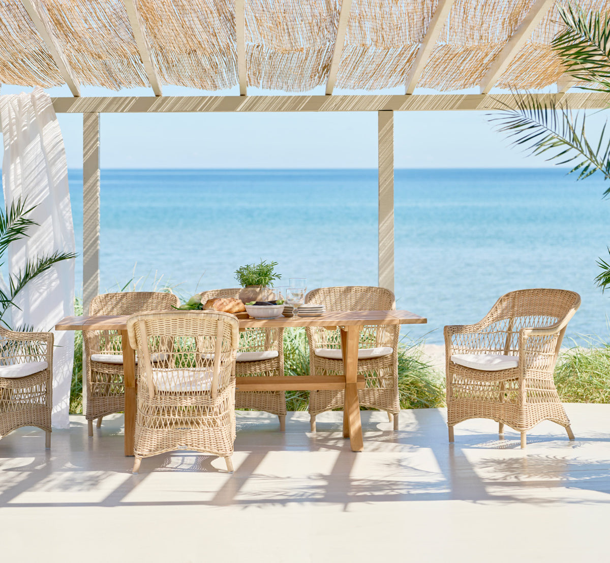 This is a lifestyle image of Colonial Table 200 in outdoor Teak, on a beachside terrace with white wooden decking and sea views.