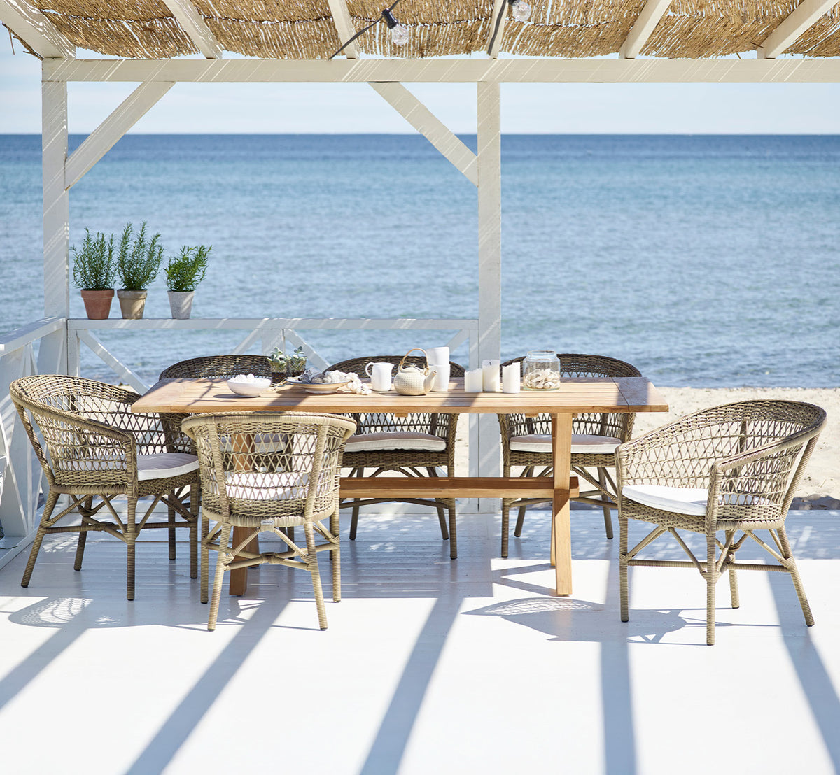 This is a lifestyle image of Colonial Table 200 in outdoor Teak, on a beachside terrace with white wooden decking and sea views.