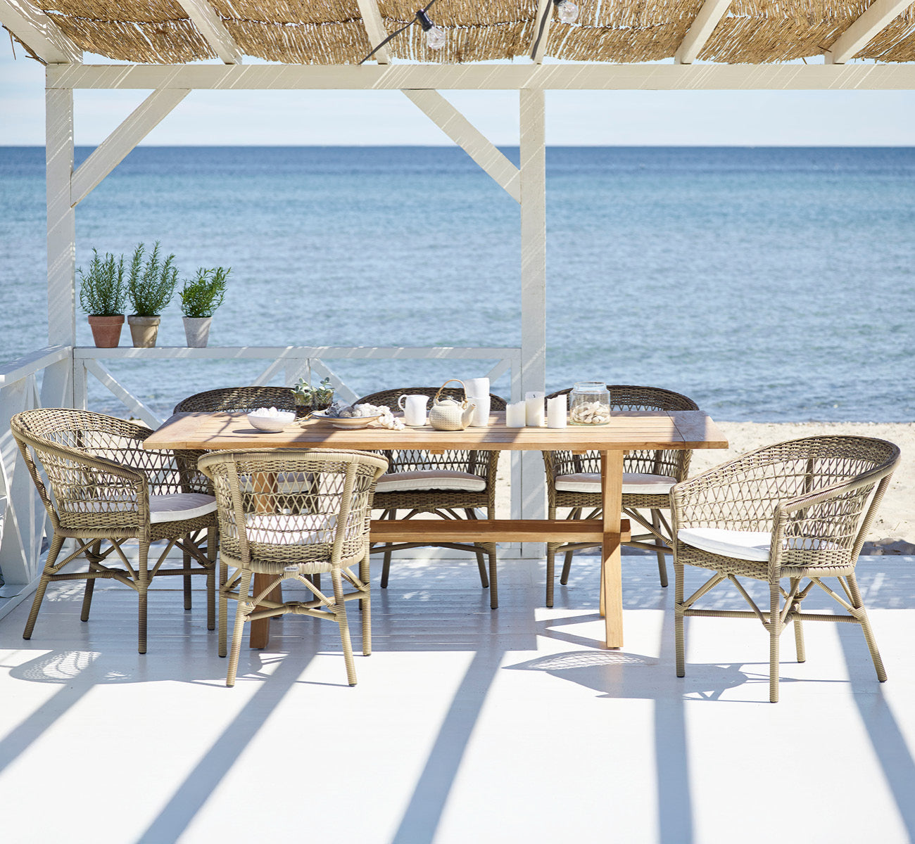 This is a lifestyle image of Colonial Table 200 in outdoor Teak, on a beachside terrace with white wooden decking and sea views.