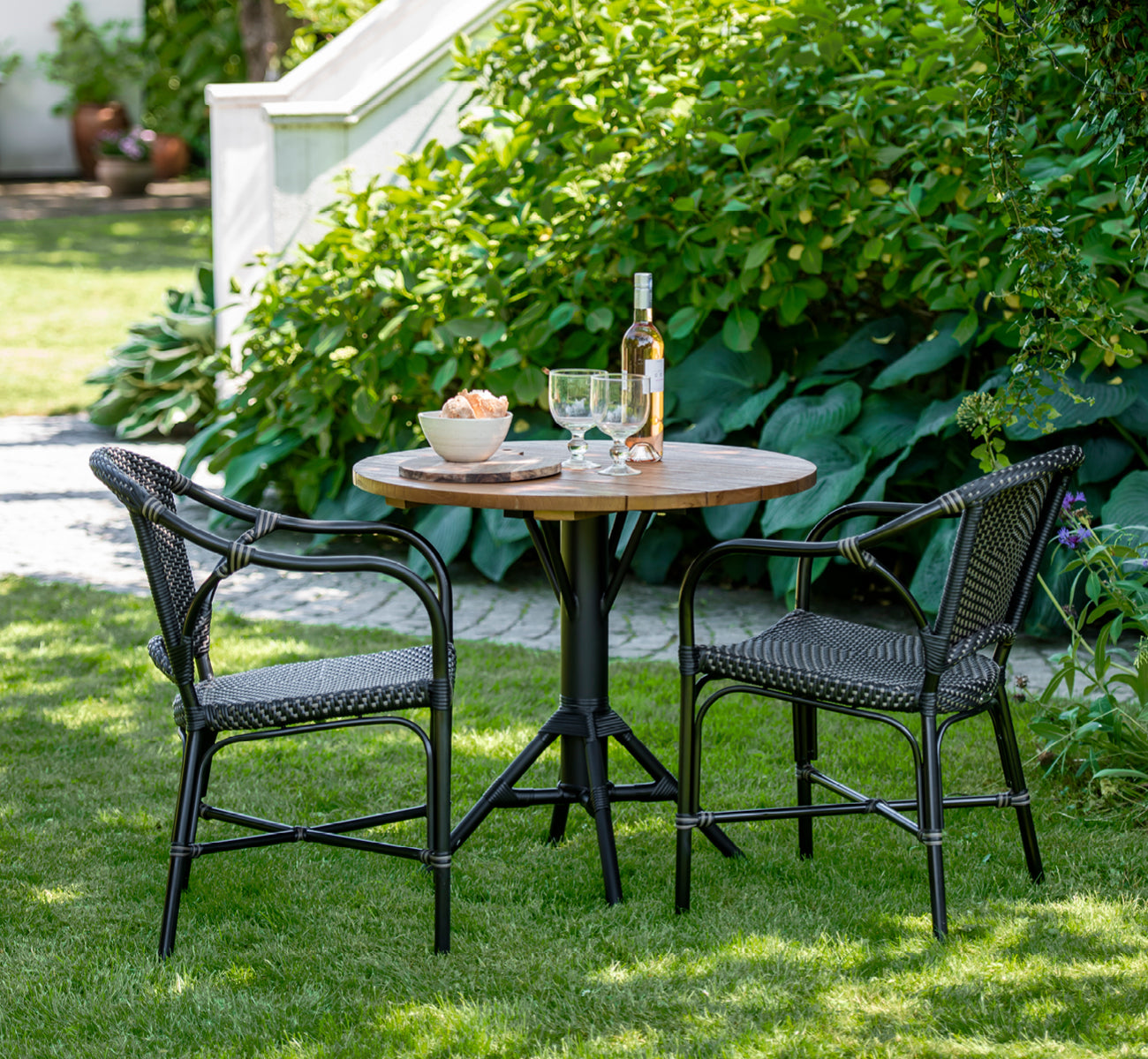 This is a lifestyle image of Nicole Cafe Table in black aluminium frame black ArtFibre. On a stone terrace with garden greenery.
