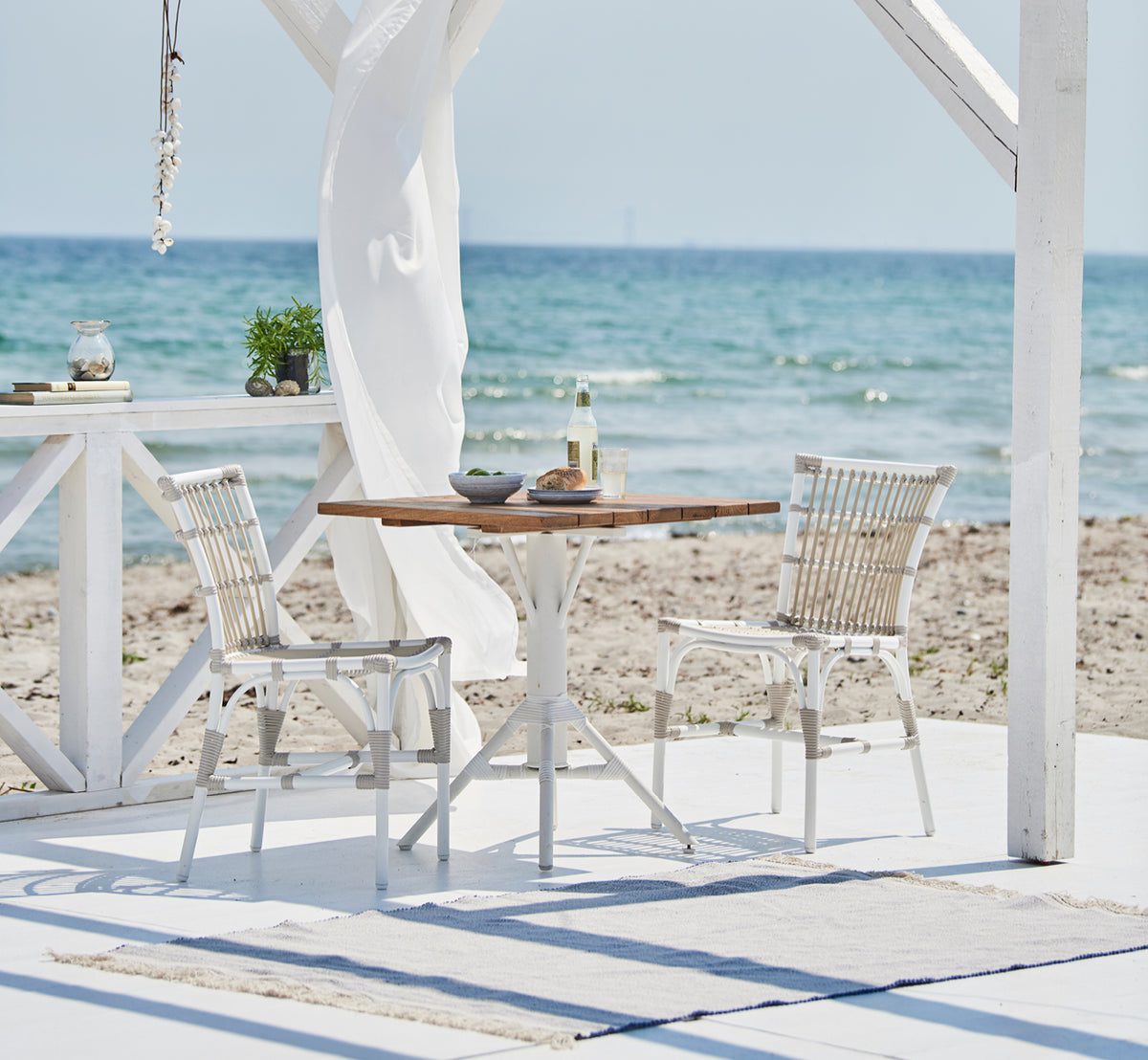 This is a lifestyle image of Nicole Cáfe Table Teak 70x70 cm in white aluminium frame white artfibre. Styled in a bright interior setting.