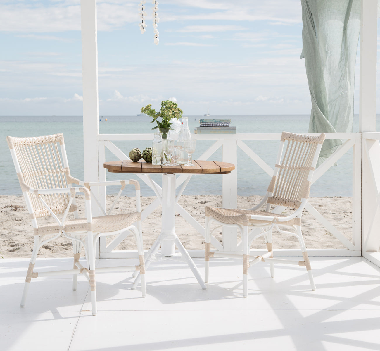 This is a lifestyle image of Nicole Cafe Table in white aluminium frame white ArtFibre. With Piano chairs on a seaside deck.
