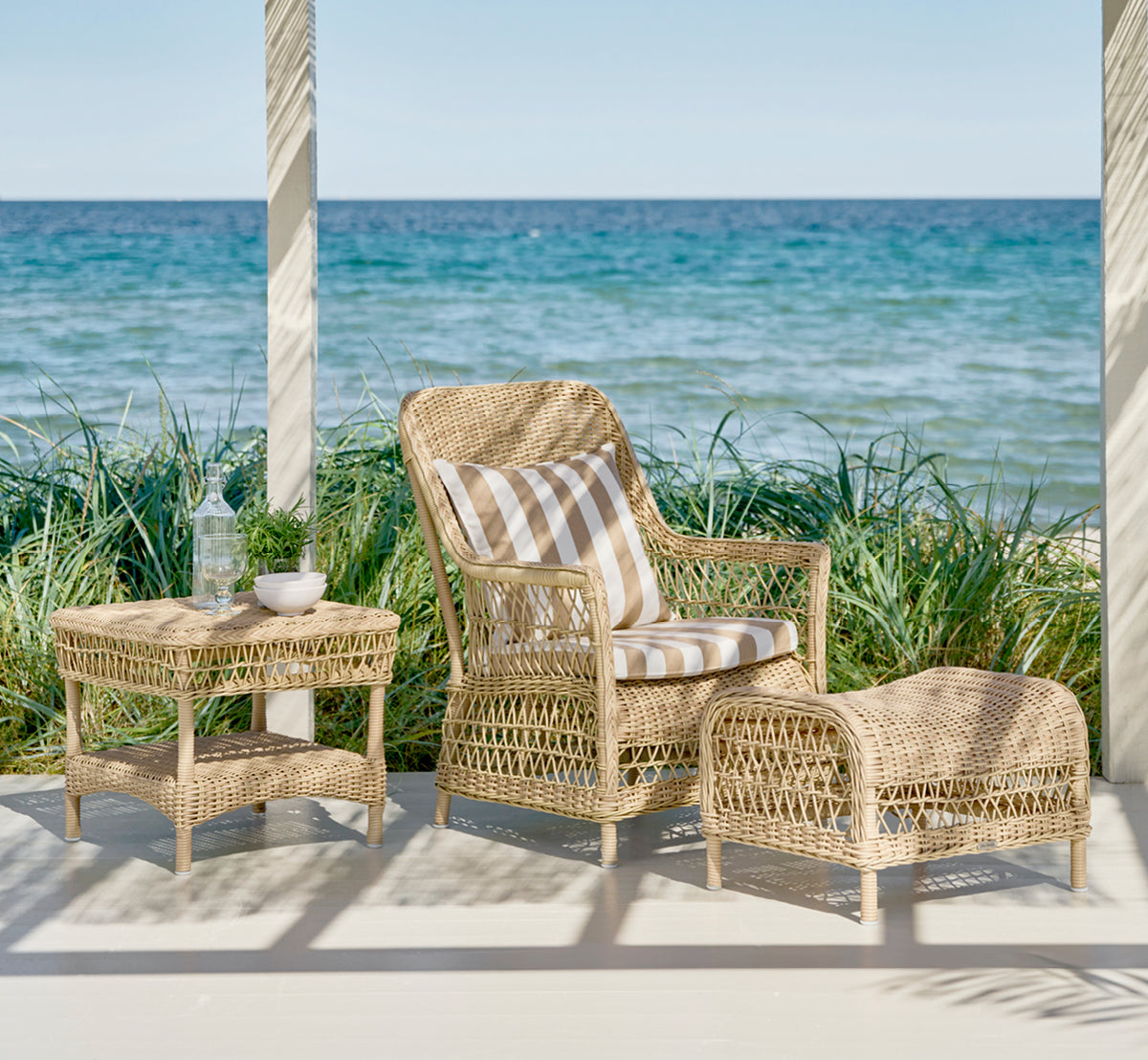 This is a lifestyle image of Susy Side Table in natural ArtFibre Aluminium, on a coastal terrace with sea views, beach grasses and white columns.