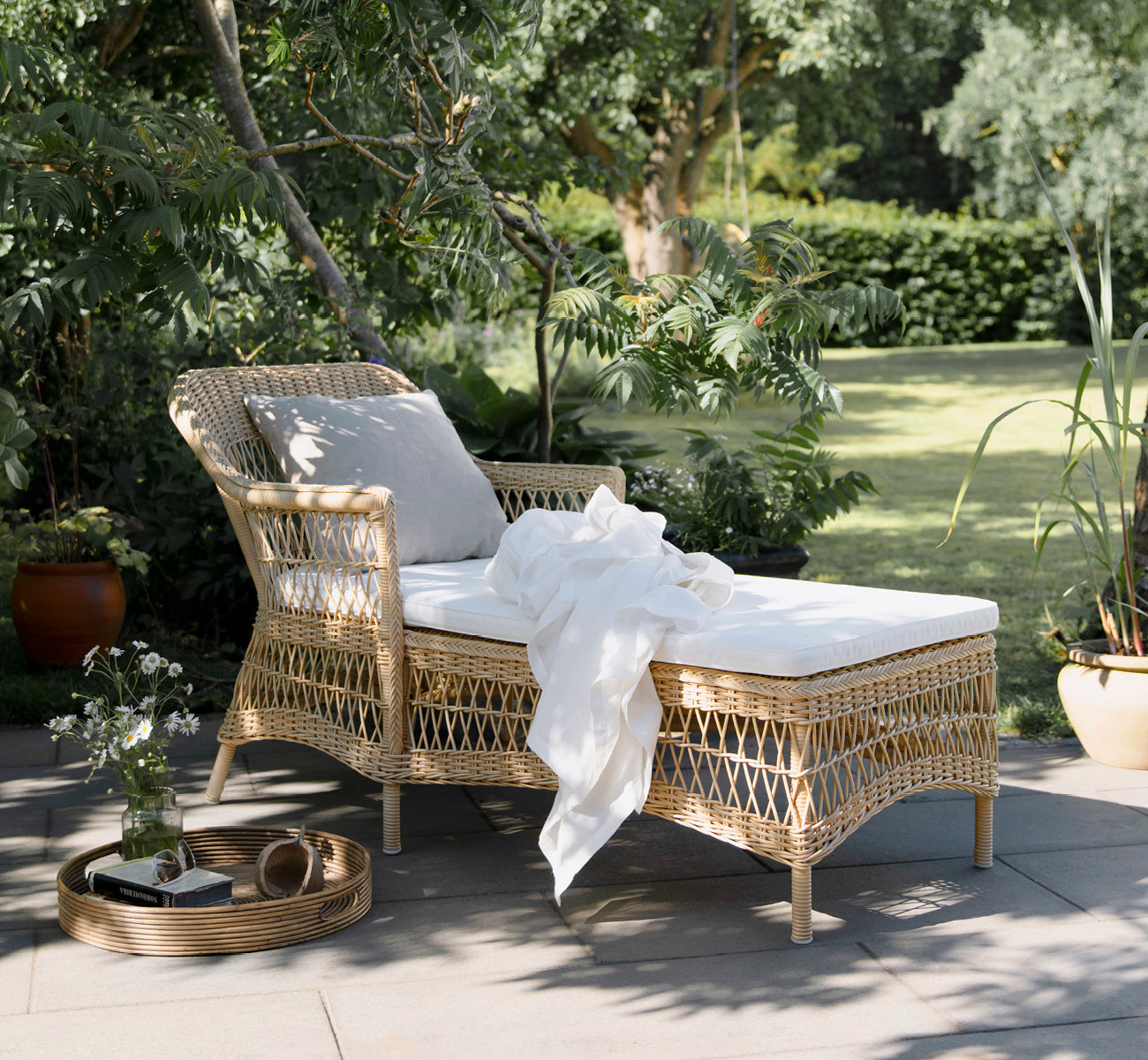 This is a lifestyle image of Olivia Exterior Sunbed in natural artfibre aluminium. In a garden setting with white throw and pillow.