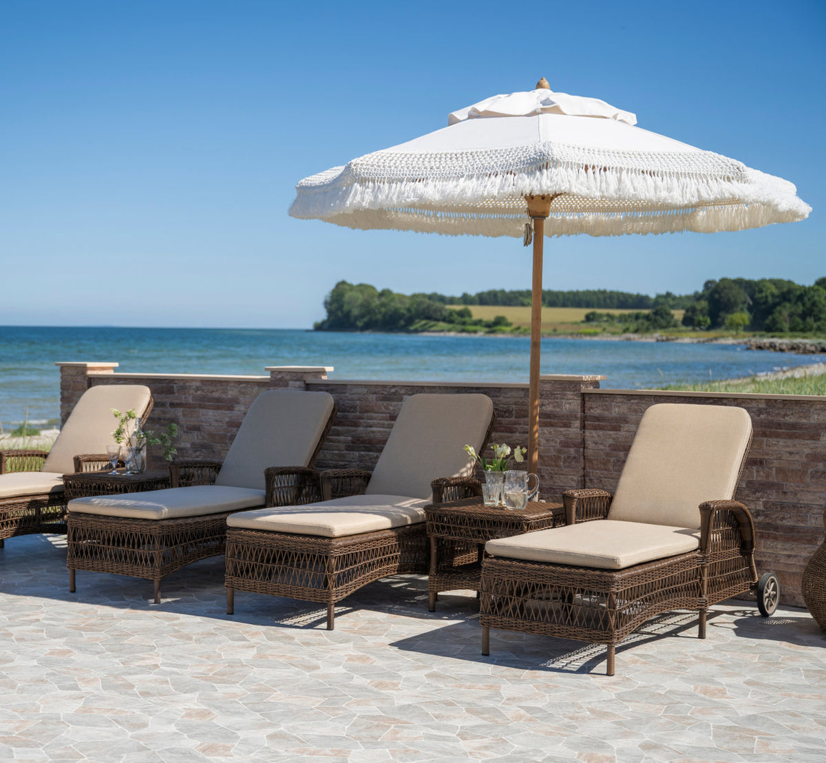 This is a lifestyle image of Daisy Exterior Sunbed in brown artfibre aluminum. Row of sunbeds under a fringed parasol by the sea.