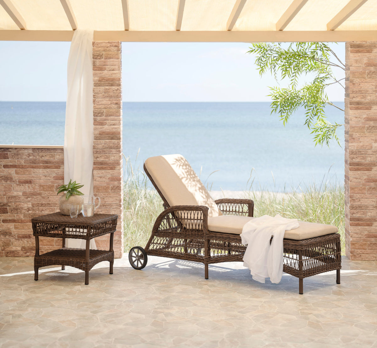 This is a lifestyle image of Daisy Exterior Sunbed in brown artfibre aluminum. With a white towel on a terrace by the sea.
