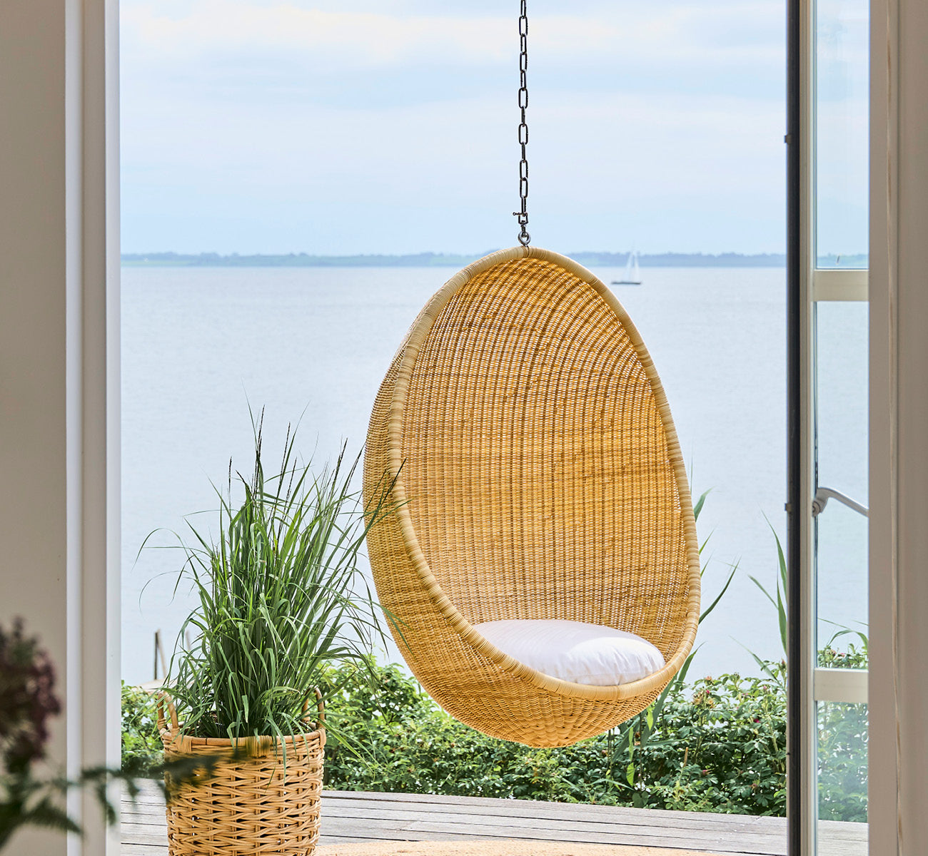 This is a lifestyle image of Chain Hanging Egg in metal, hung by a glass door with water views and surrounding green plants.