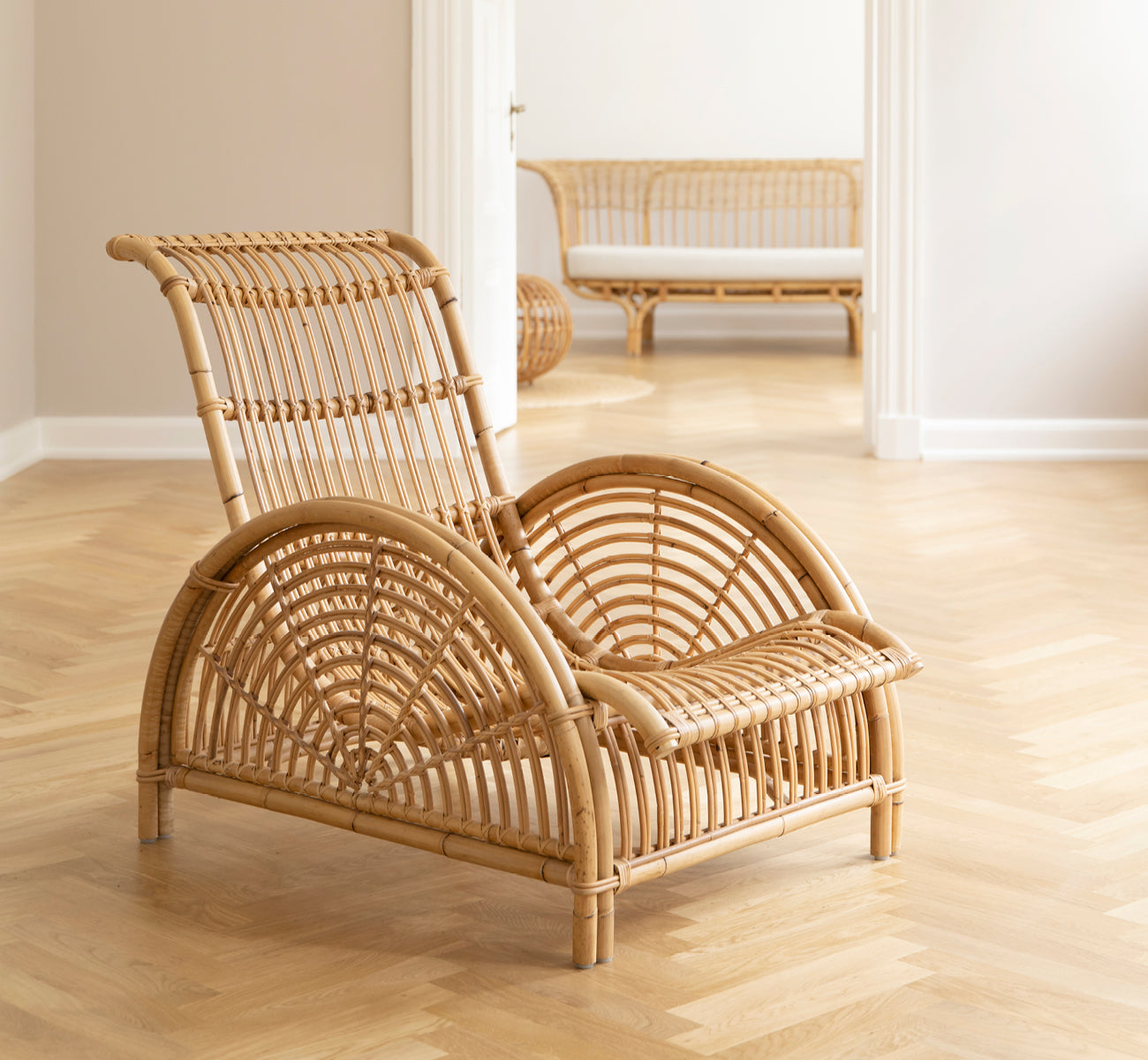 This is a lifestyle image of Paris Lounge Chair in polished natural rattan. Placed on a herringbone parquet floor beside a matching bench.