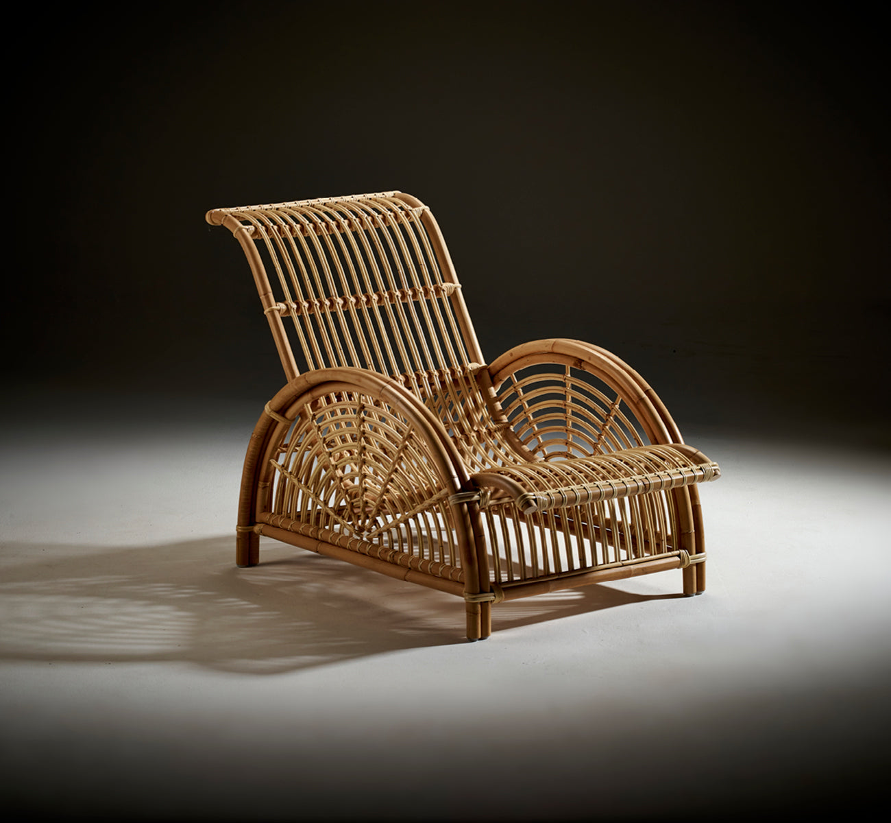 This is a lifestyle image of Paris Lounge Chair in polished natural rattan. Placed on a herringbone parquet floor beside a matching bench.