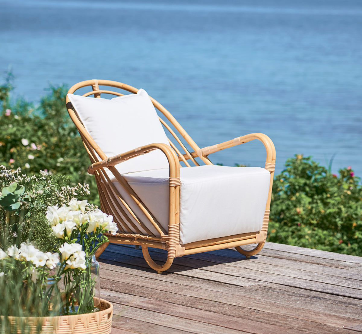 This is a lifestyle image of Charlottenborg Exterior Lounge Chair in natural Aluminium ArtFibre, on an outdoor wooden deck with flowers and sea views.