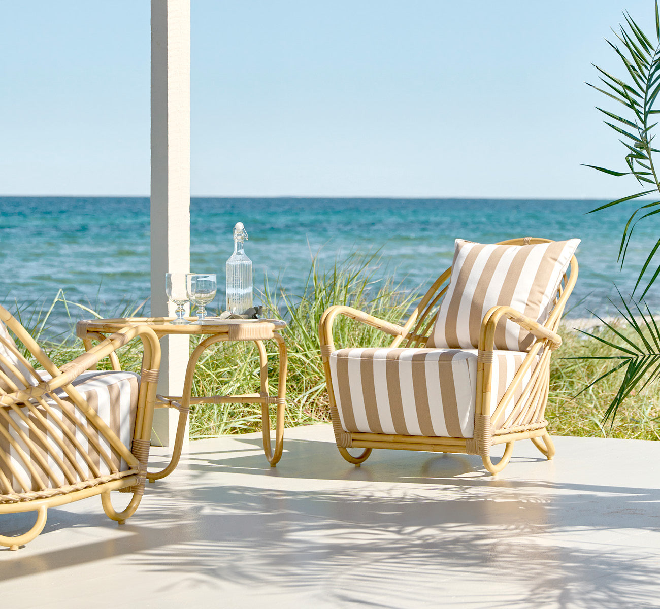 This is a lifestyle image of Charlottenborg Exterior Lounge Chair in natural Aluminium ArtFibre, on an outdoor wooden deck with flowers and sea views.