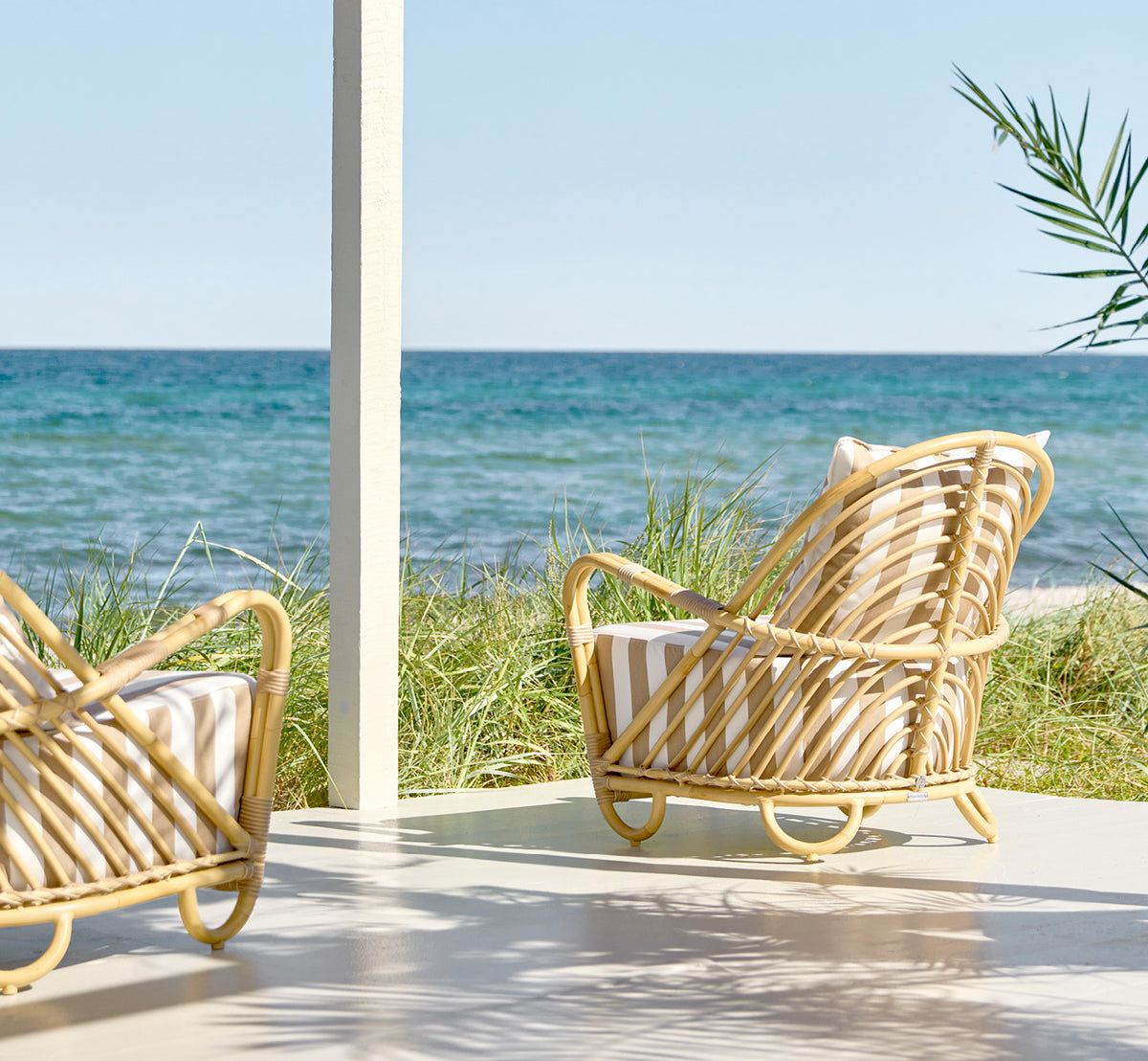 This is a lifestyle image of Charlottenborg Exterior Lounge Chair in natural Aluminium ArtFibre, on an outdoor wooden deck with flowers and sea views.