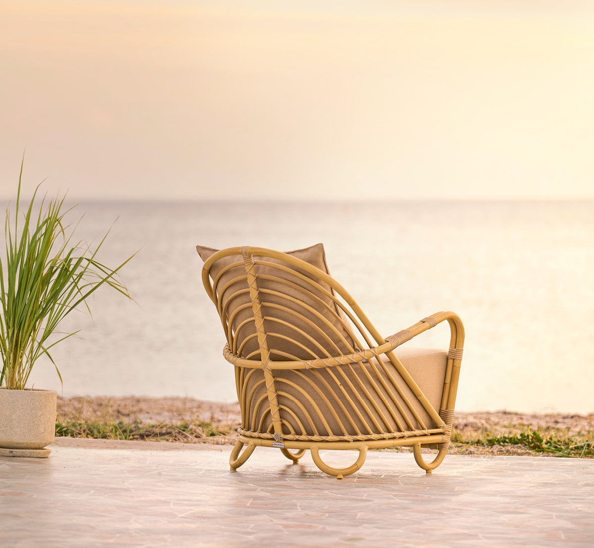 This is a lifestyle image of Charlottenborg Exterior Lounge Chair in natural Aluminium ArtFibre, on an outdoor wooden deck with flowers and sea views.