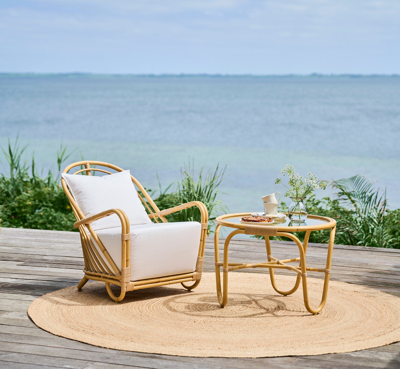 This is a lifestyle image of Charlottenborg Exterior Lounge Chair in natural Aluminium ArtFibre, on an outdoor wooden deck with flowers and sea views.