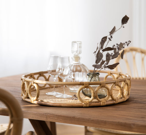 This is a lifestyle image of Mira Tray in natural rattan. On a dining table with glassware and dried eucalyptus in a bright room.