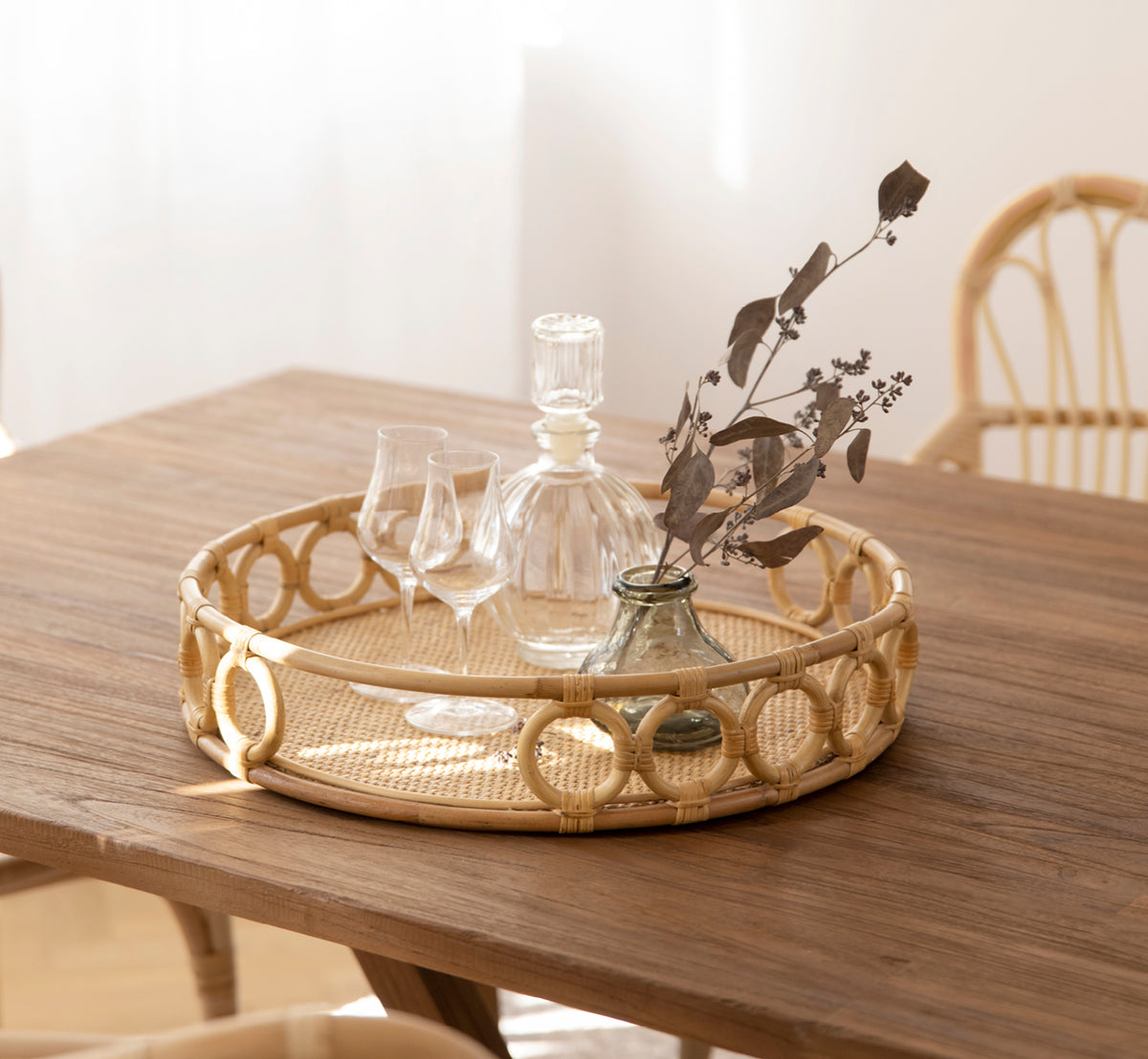 This is a lifestyle image of Mira Tray in natural rattan. On a teak table holding crystal glasses and a vase with dried branches.