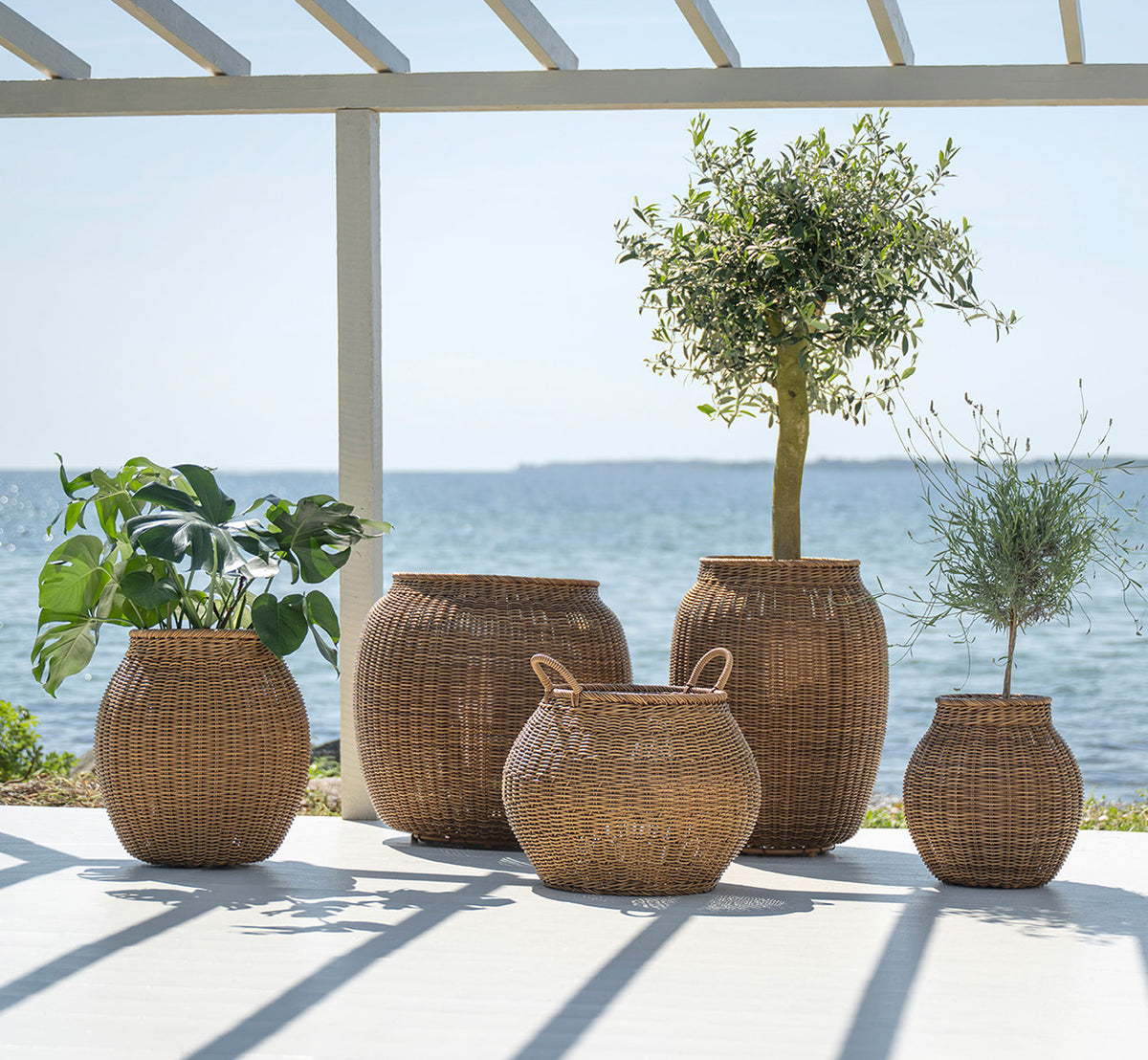 This is a lifestyle image of Alma Basket in chestnut core weave ArtFibre Aluminium, outdoors on a sun-lit patio with a white wall and tile flooring.