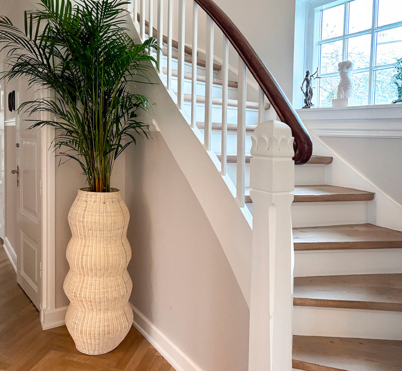 This is a lifestyle image of Unda Basket H102 cm in natural core weave iron frame. Used as a tall planter next to a curved staircase in a bright hall.