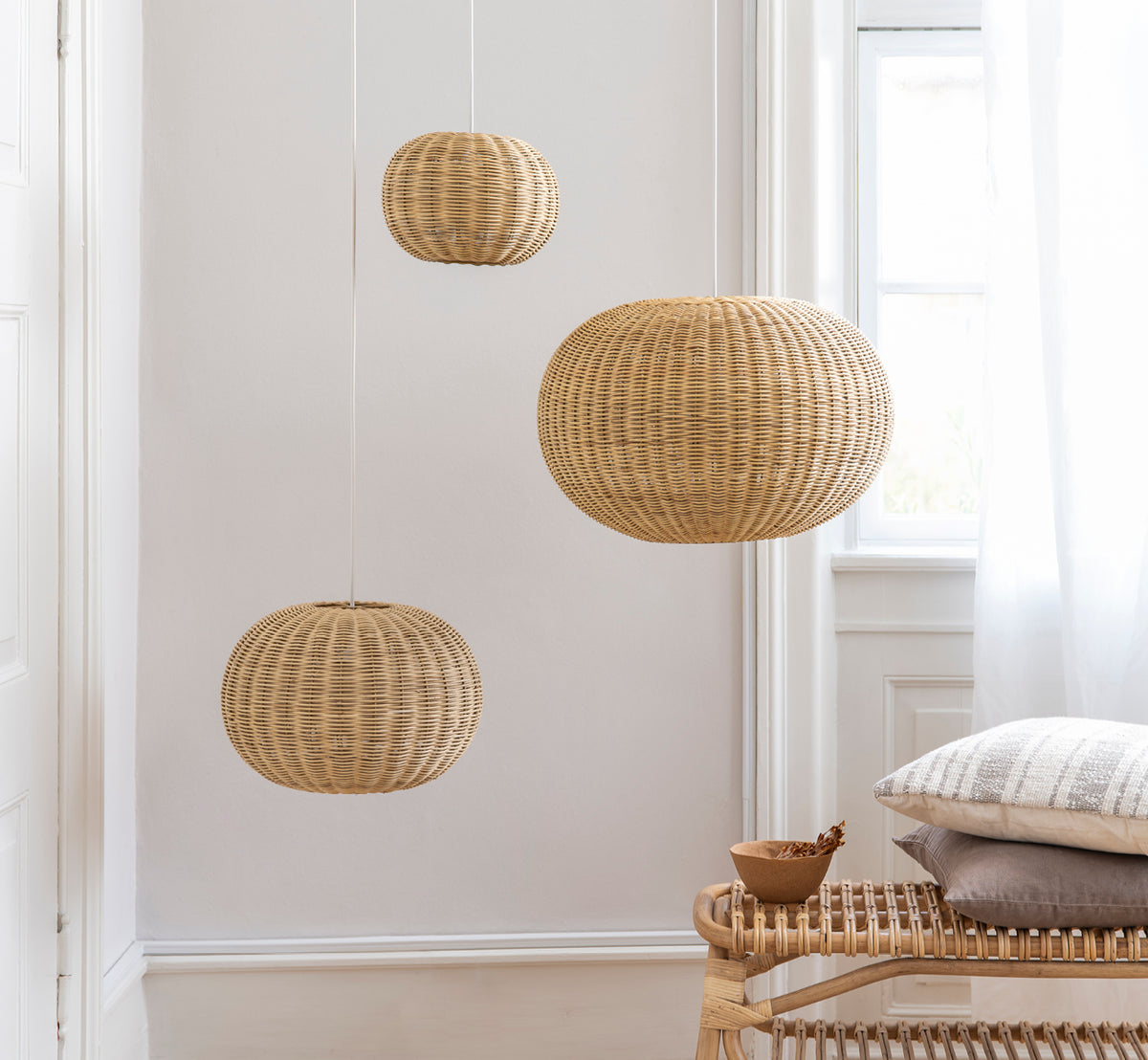 This is a lifestyle image of Tangelo Lampshade Ø49 cm | Medium in natural rattan. Three pendants hanging against a white wall above a rattan bench.