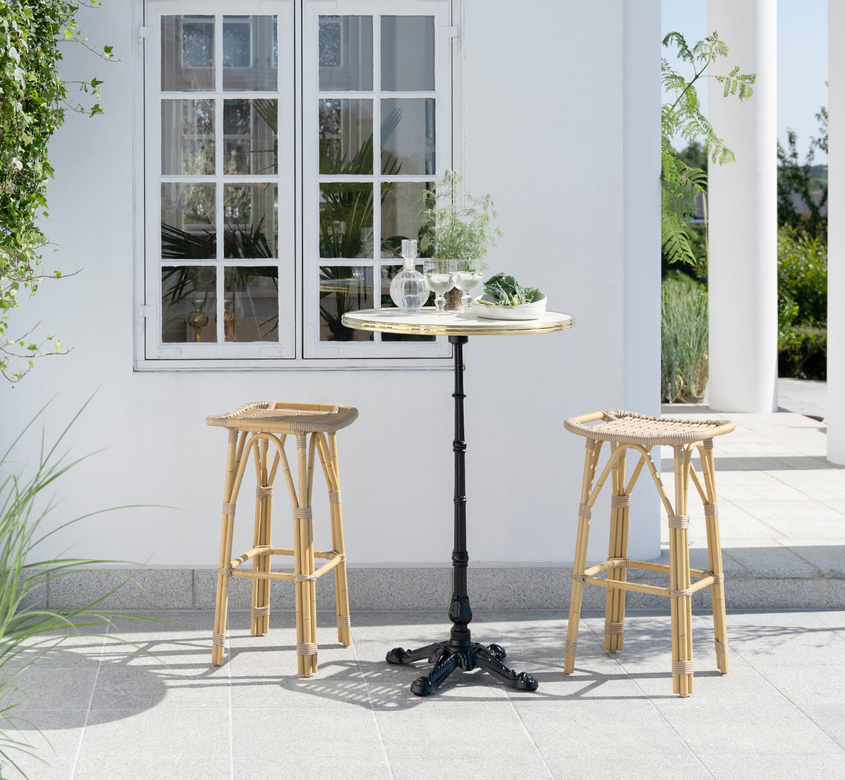 This is a lifestyle image of Lyon Bar Table Versalite Marble Ø70 cm in cast iron. Elegant patio with bar stools and green plants.