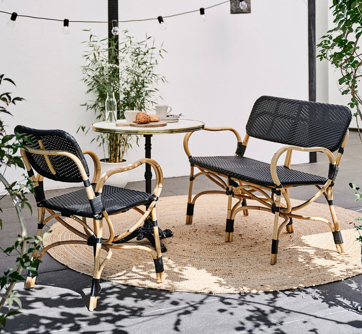 This is a lifestyle image of Bistro Bench in skin on Rattan frame, in an outdoor café setting with a bistro table on dark tile flooring.