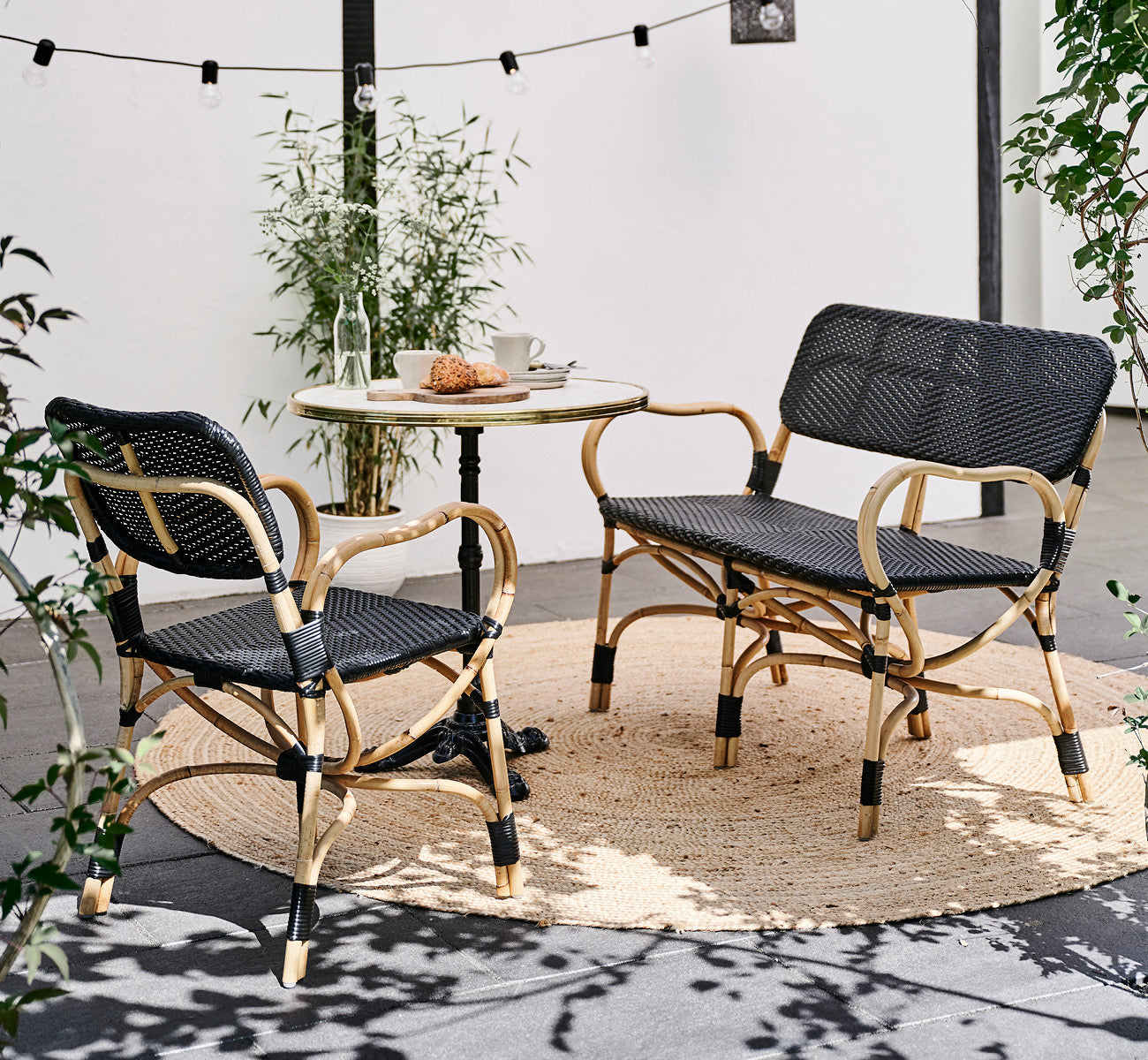 This is a lifestyle image of Bistro Bench in skin on Rattan frame, in an outdoor café setting with a bistro table on dark tile flooring.