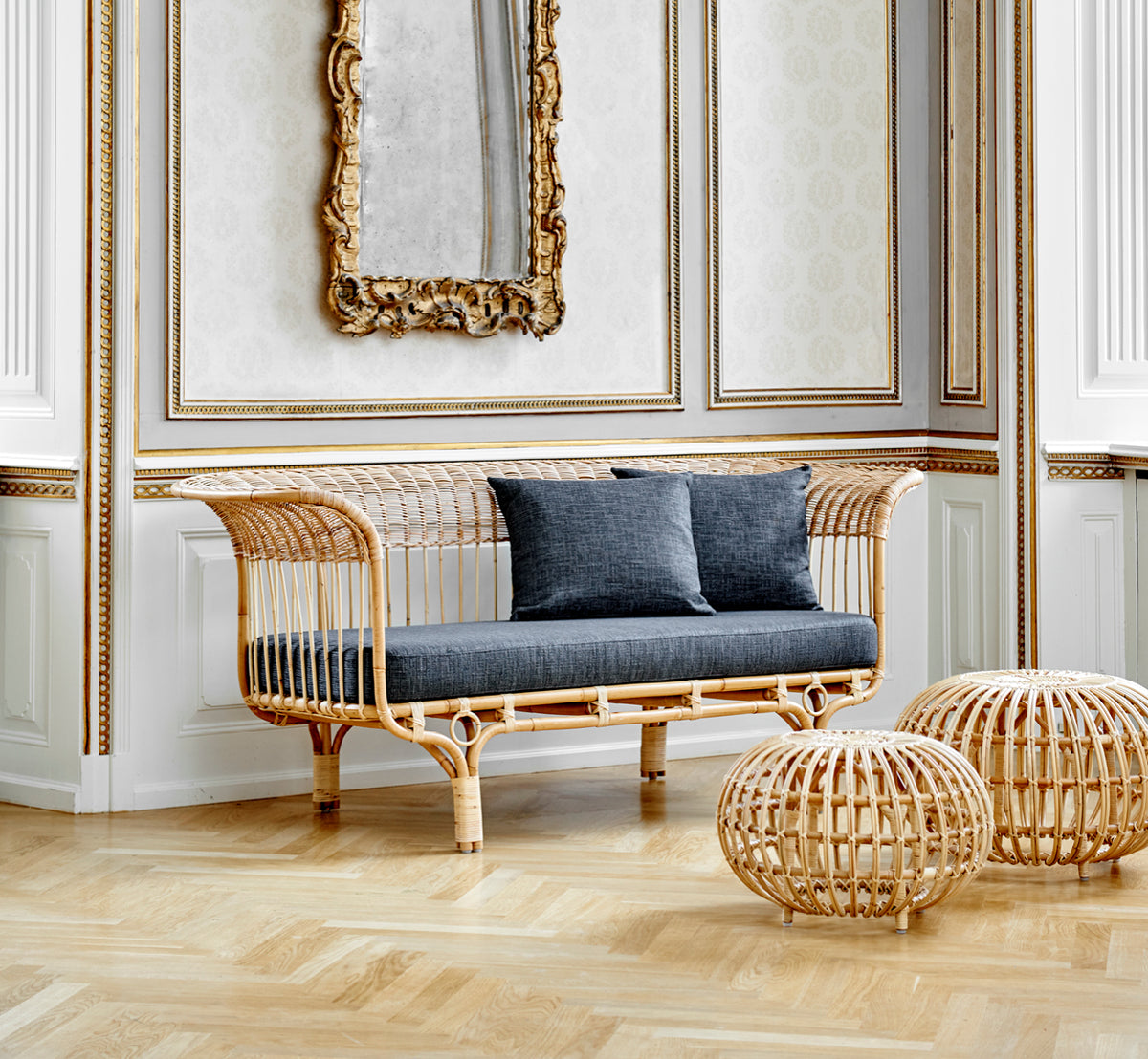 This is a lifestyle image of Ottoman Large in natural skin on rattan. Clustered in a classical panelled interior with a gilded clock.