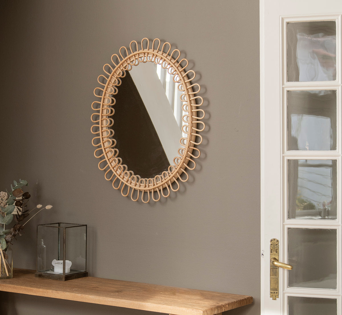 This is a lifestyle image of Luella Mirror in polished natural rattan. Hanging on a gray wall above a wooden shelf with eucalyptus and glass décor.