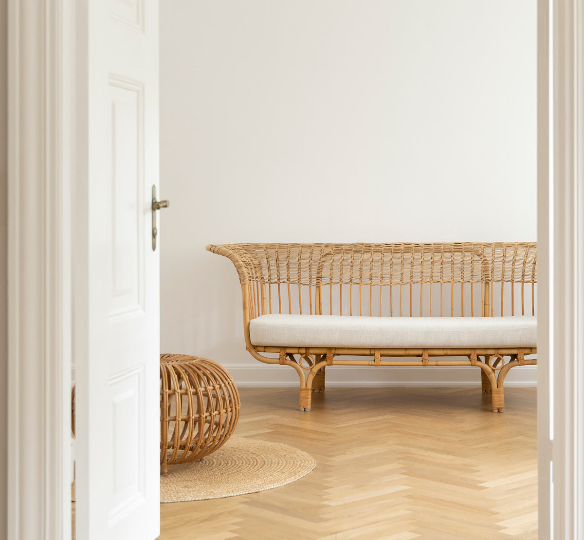 This is a lifestyle image of Belladonna Sofa in polished natural rattan, in a bright Scandinavian interior seen through a white doorway.