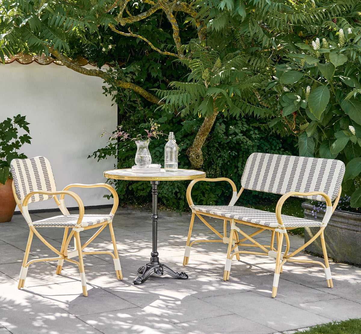 This is a lifestyle image of Bistro Exterior Chair in natural Aluminium ArtFibre, in an outdoor garden café with a bistro table and surrounding trees.