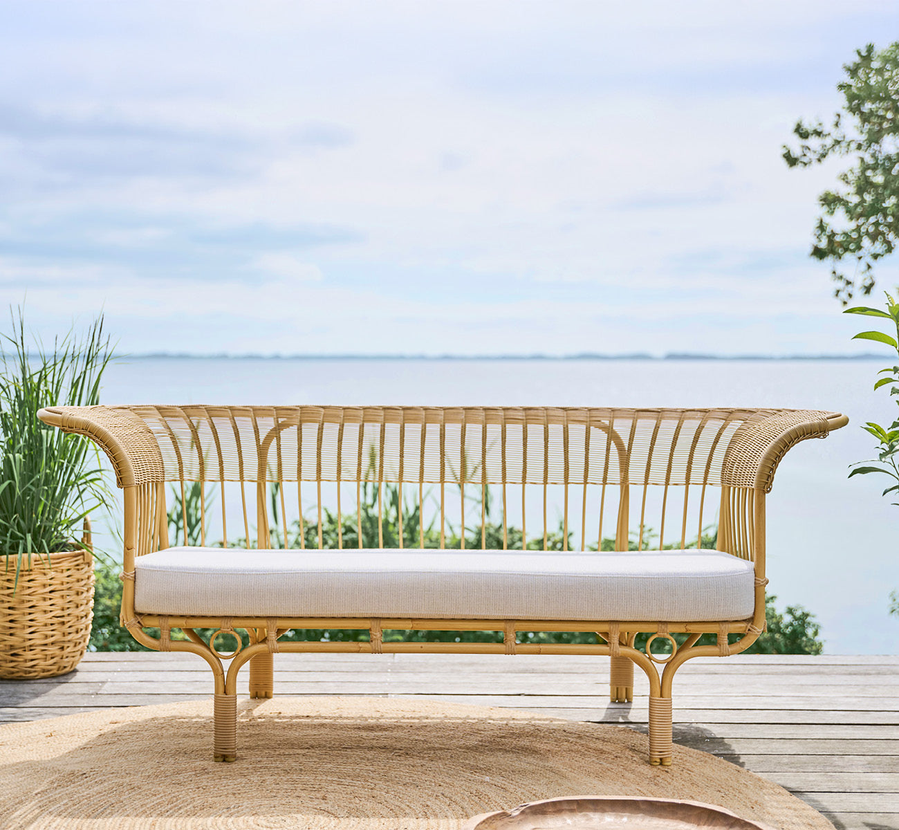 This is a lifestyle image of Belladonna Exterior Sofa in natural Aluminium ArtFibre, on an outdoor wooden deck overlooking the water.