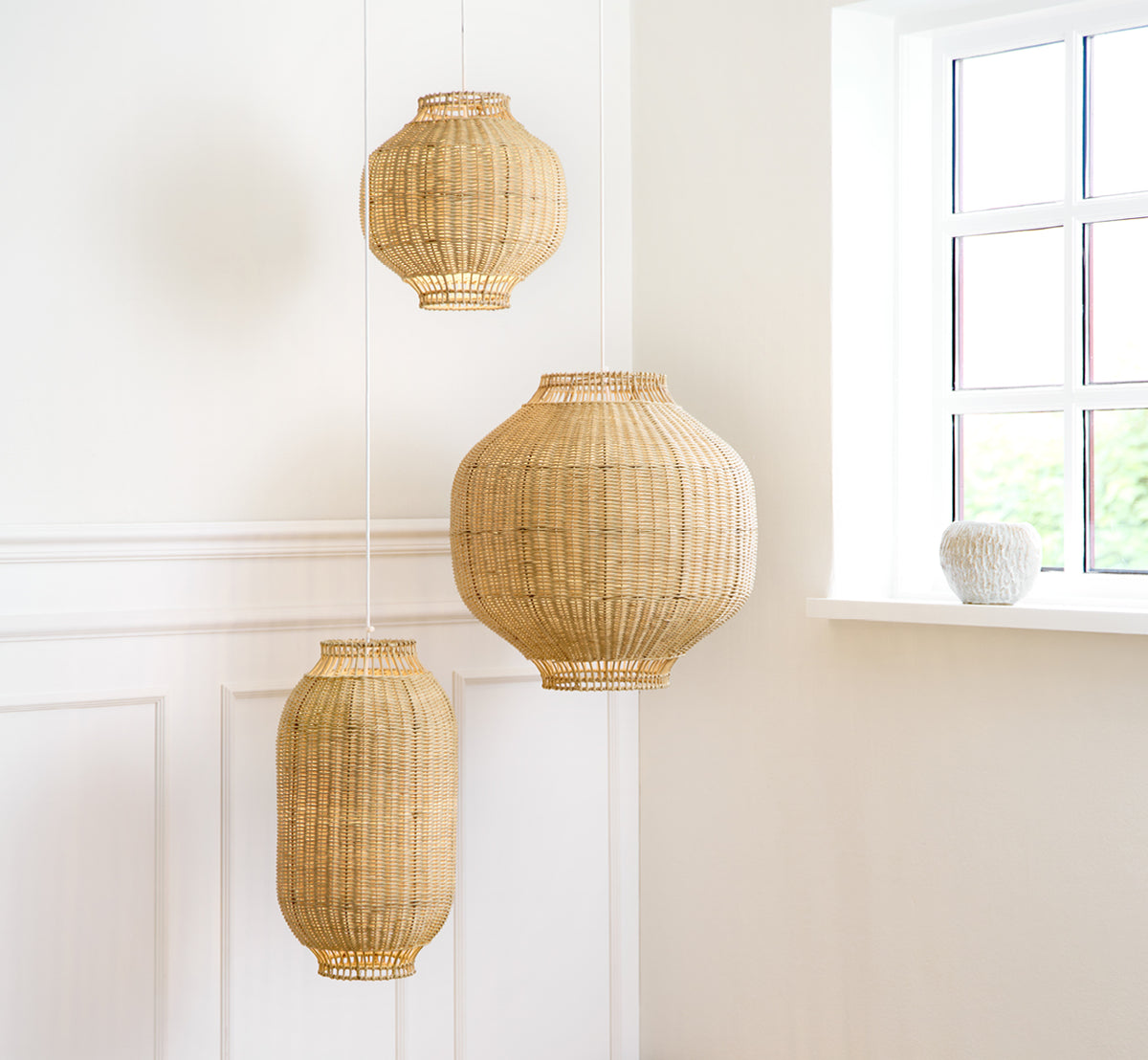 This is a lifestyle image of Kou Lampshade in natural core weave rattan frame. Three sizes hang together in a bright white paneled room.