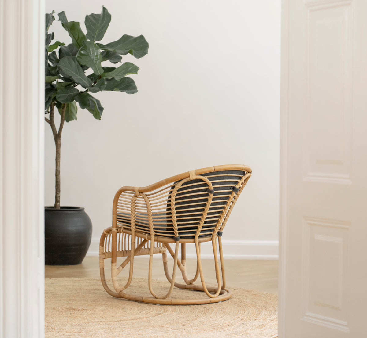 This is a lifestyle image of Swing Lounge Chair in polished natural rattan. Standing alone beside a leafy plant on a herringbone parquet floor.