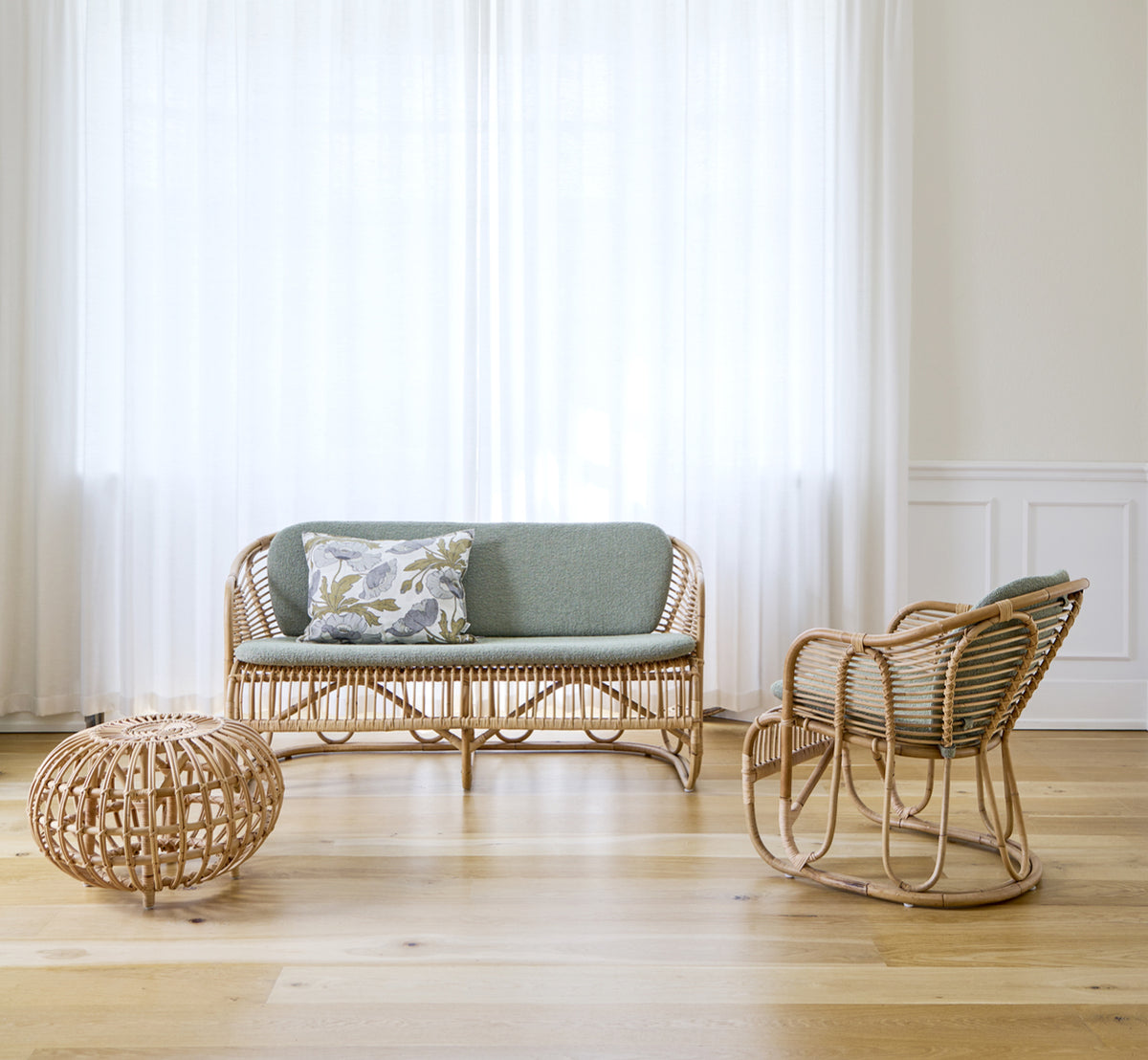 This is a lifestyle image of Swing Lounge Chair in polished natural rattan. Standing alone beside a leafy plant on a herringbone parquet floor.