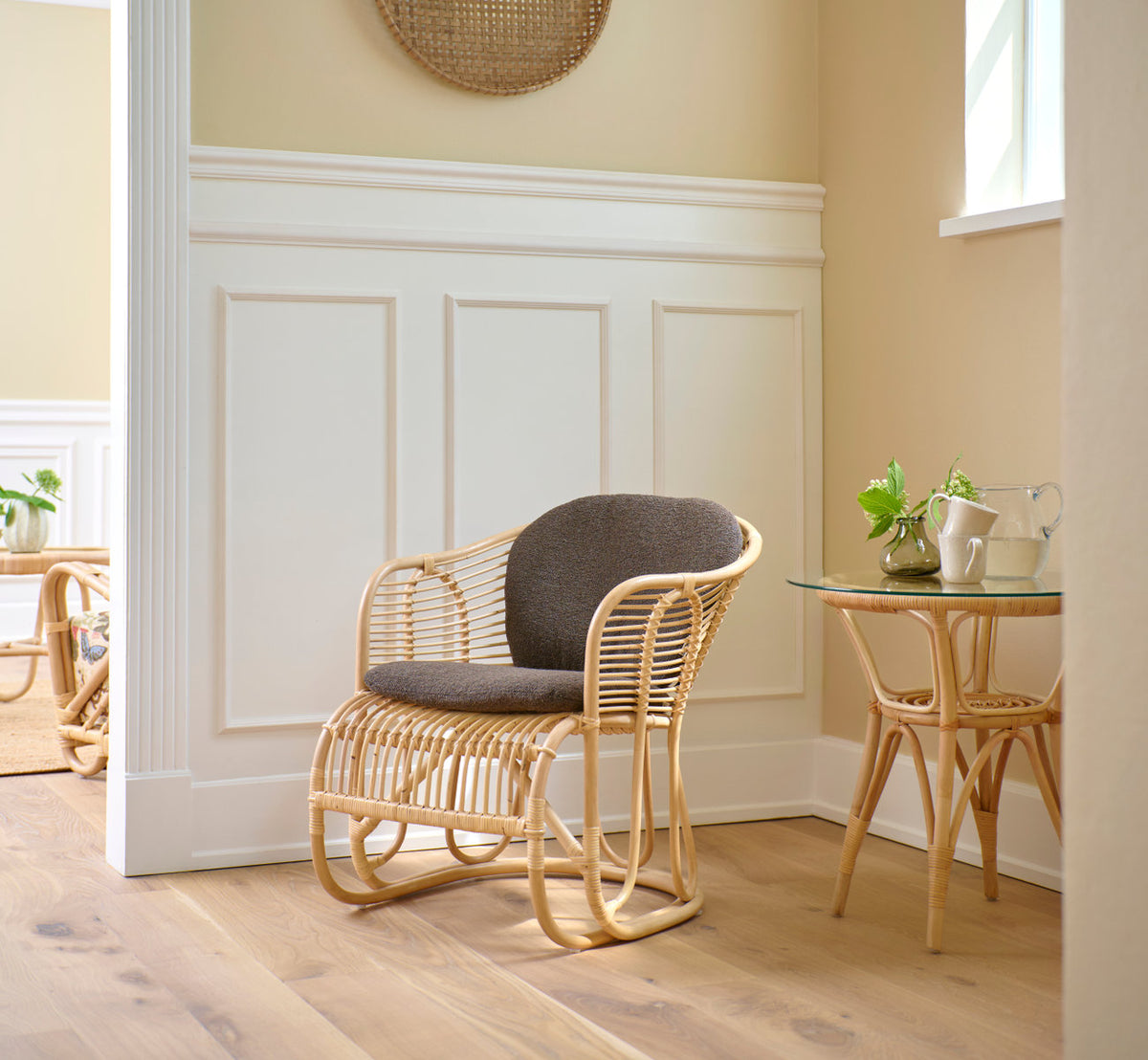 This is a lifestyle image of Swing Lounge Chair in polished natural rattan. With grey cushion in a bright paneled corner.