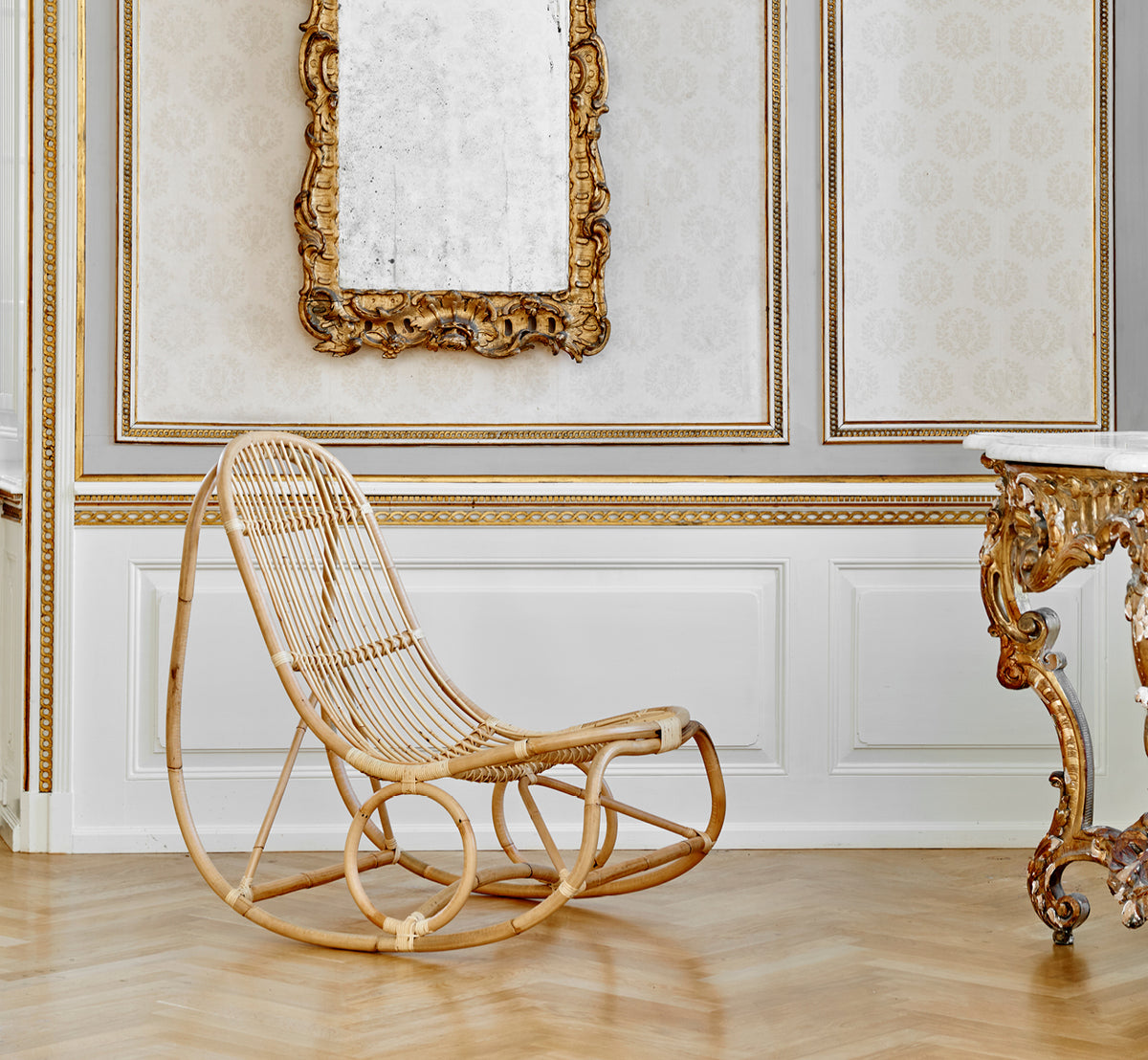 This is a lifestyle image of Nanny Rocking Chair in polished natural rattan. In an ornate gilded room with baroque mirror and console.