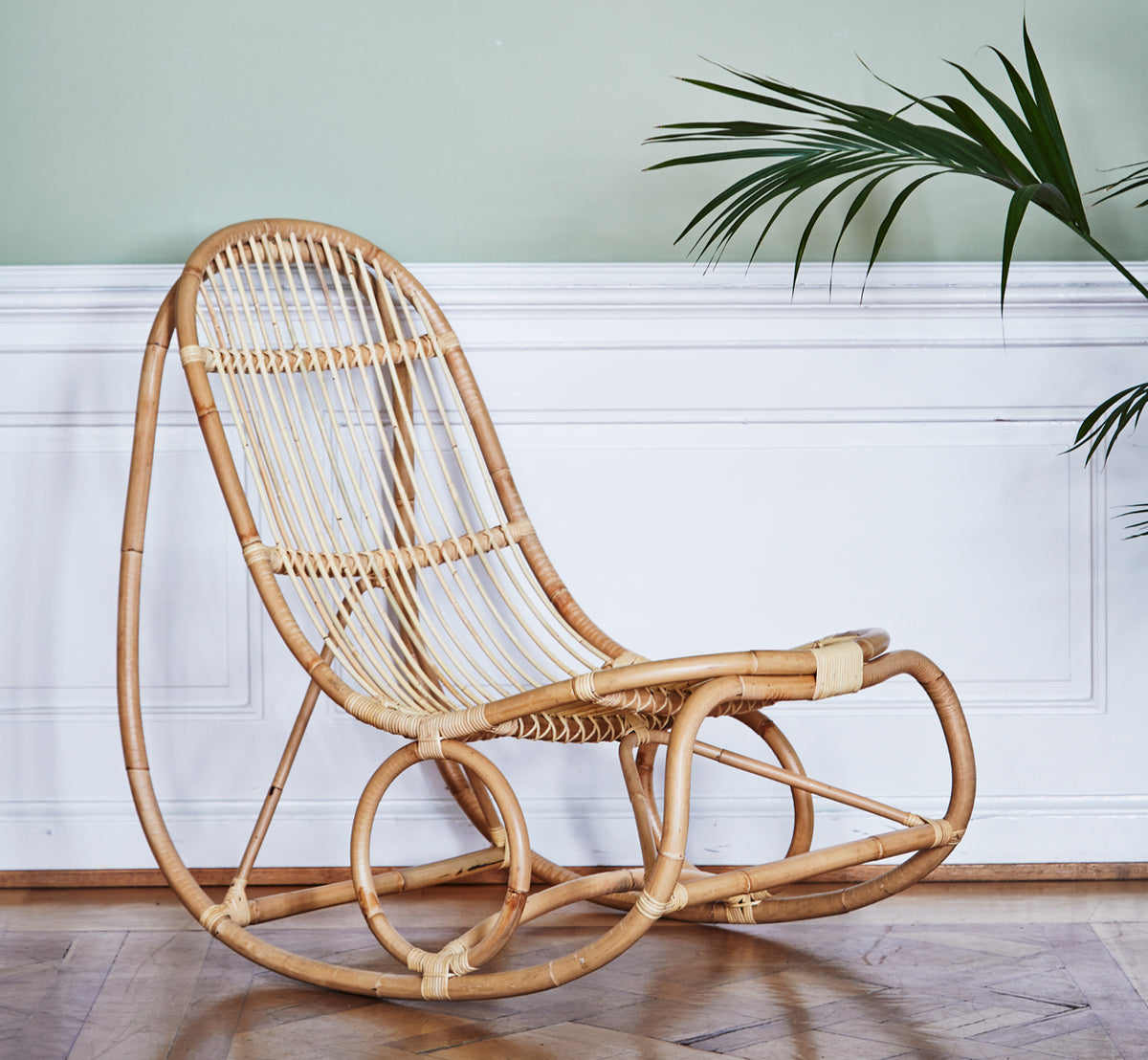 This is a lifestyle image of Nanny Rocking Chair in polished natural rattan. Against a sage green wall with white paneling and palm.