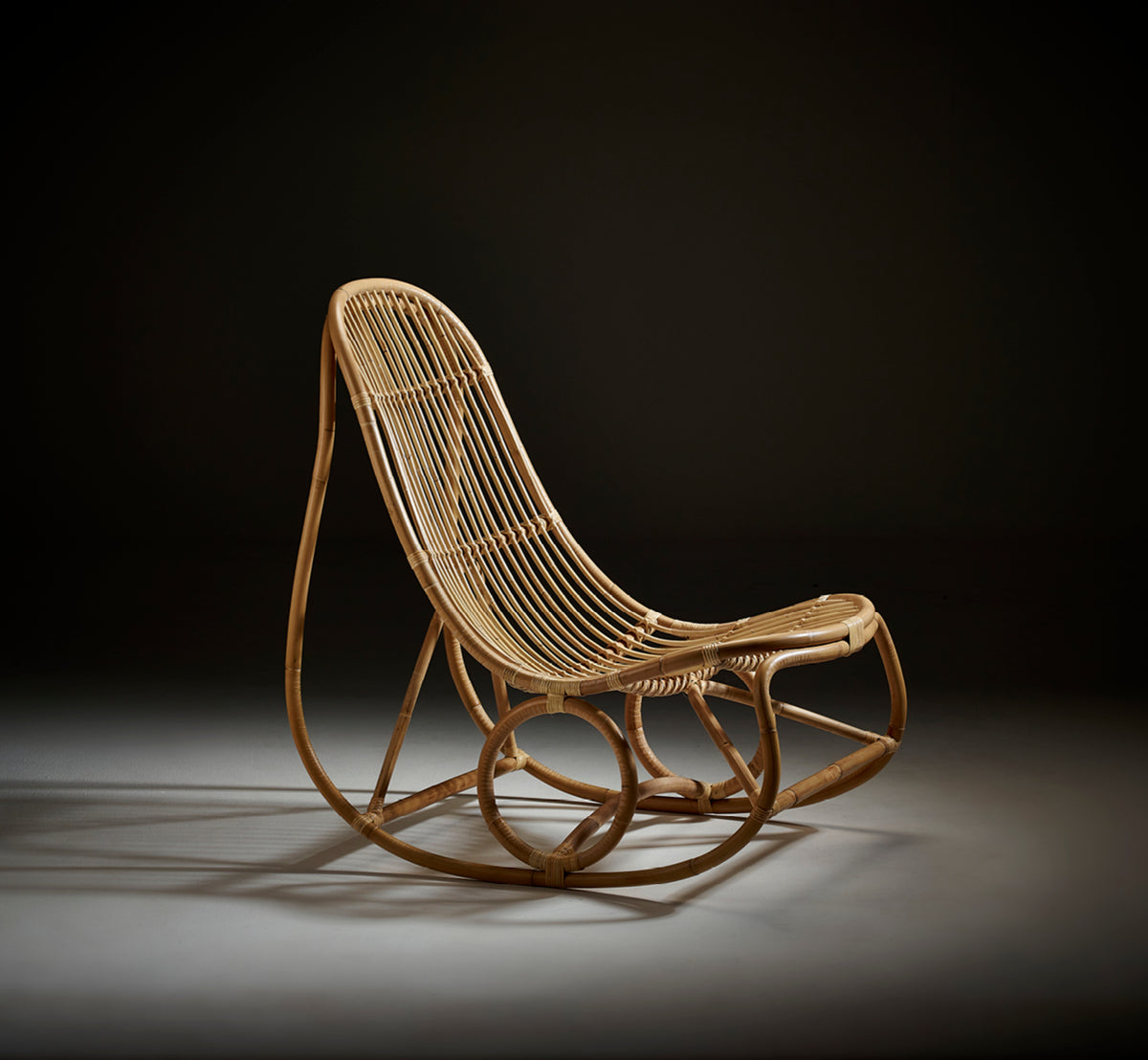 This is a lifestyle image of Nanny Rocking Chair in polished natural rattan. Studio shot on dark background highlighting its curves.