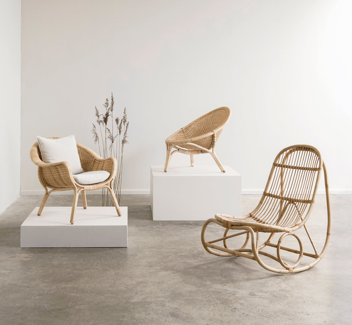 This is a lifestyle image of Nanny Rocking Chair in polished natural rattan. In a gallery with rattan chairs on white display plinths.
