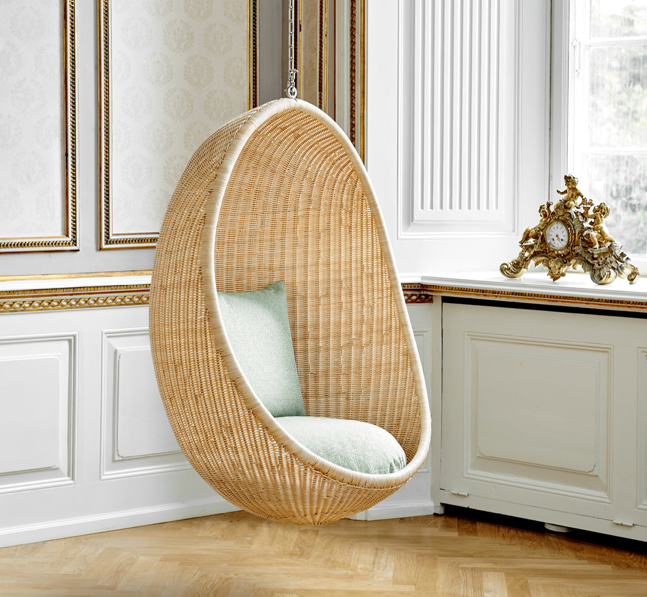 This is a lifestyle image of Hanging Egg Chair in natural core weave Rattan frame, in an elegant classical interior with ornate golden mouldings.