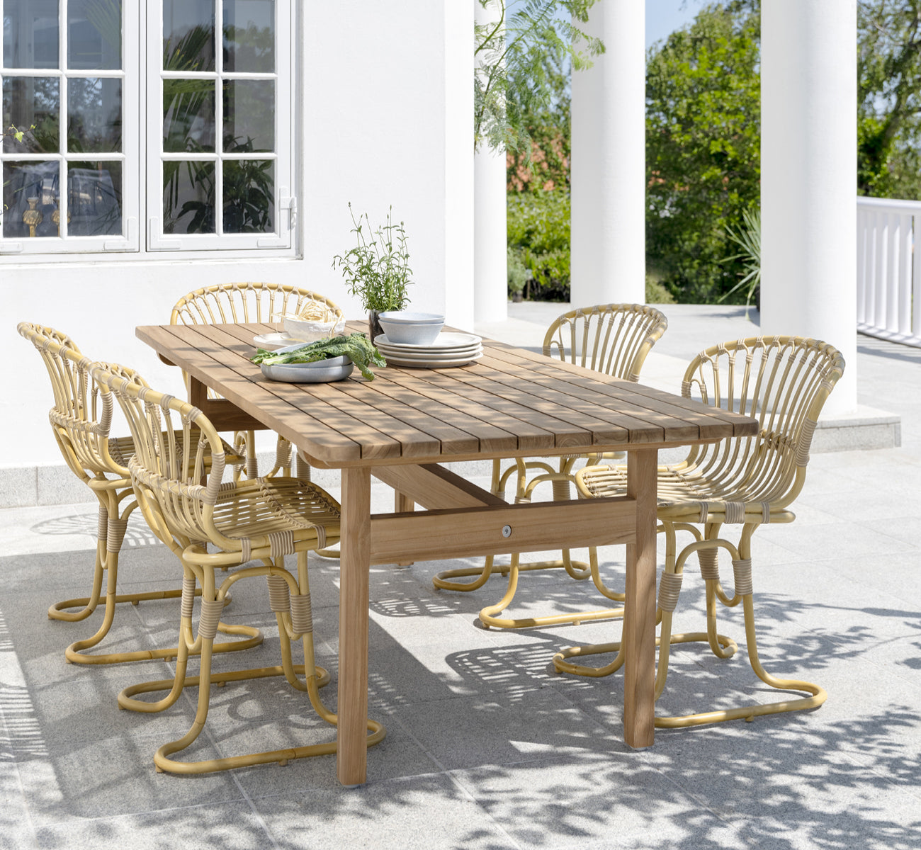 This is a lifestyle image of Rita Exterior Dining Chair in natural aluminium artfibre. Paired with a bistro table on a white colonnaded terrace.
