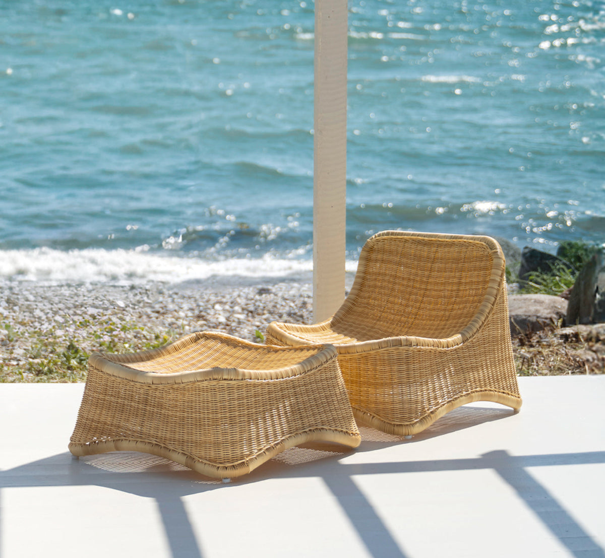 This is a lifestyle image of Chill Lounge Chair in natural core weave ArtFibre Aluminium, on a waterside wooden deck with coastal greenery and sea.