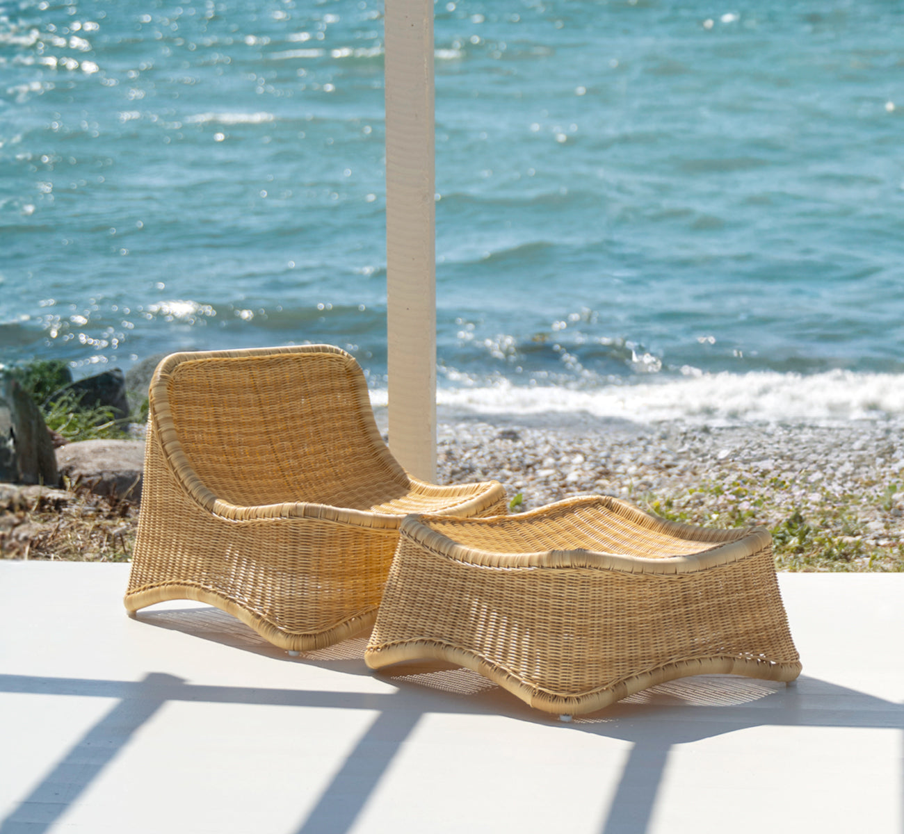 This is a lifestyle image of Chill Lounge Chair in natural core weave ArtFibre Aluminium, on a waterside wooden deck with coastal greenery and sea.