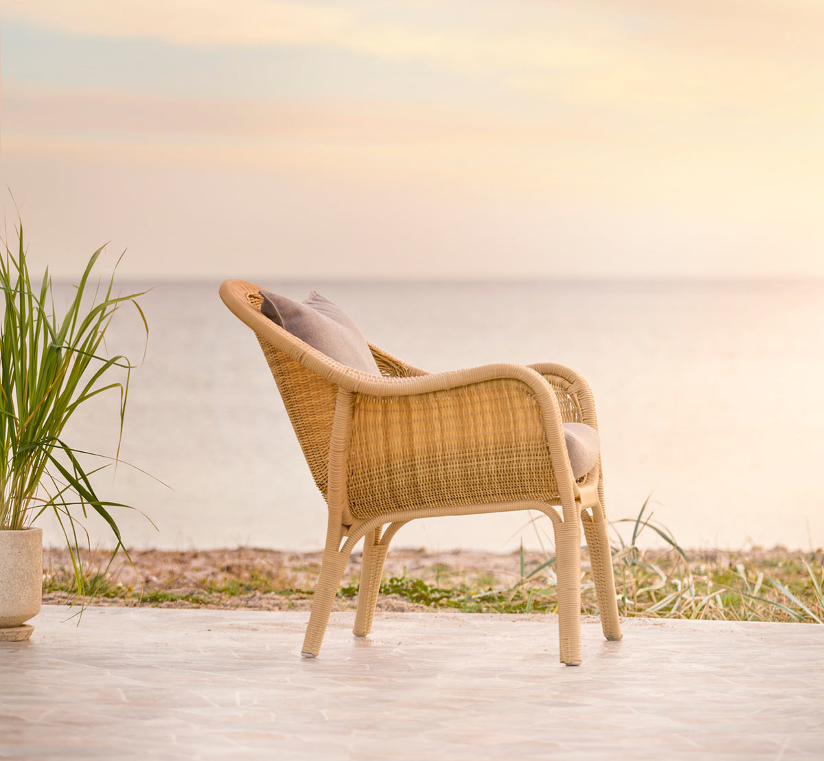 This is a lifestyle image of Madame Lounge Chair in natural core weave ArtFibre Aluminium, on an open coastal terrace at sunset with sea views and.